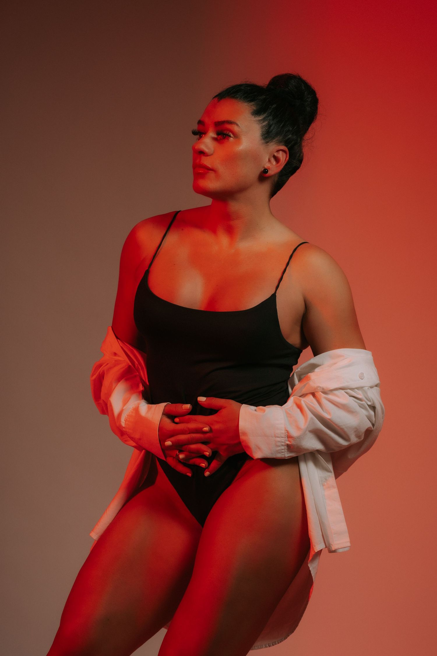 Athletic woman in black bodysuit with white jacket poses under dramatic red studio lighting.
