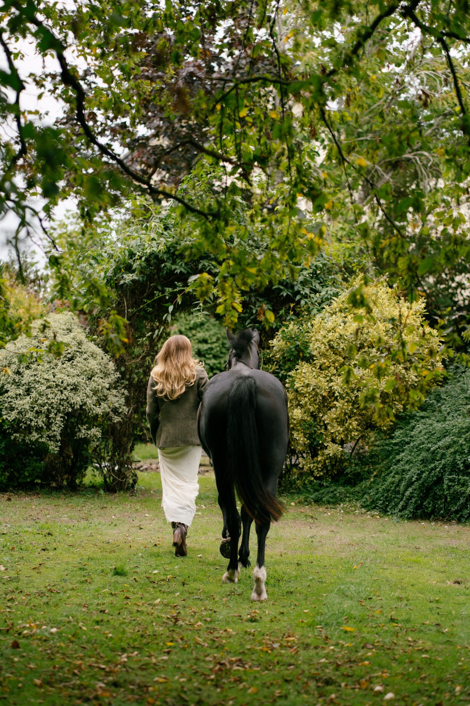 Woman with blonde hair walks alongside a dark horse through a lush green garden with trees and shrubs.