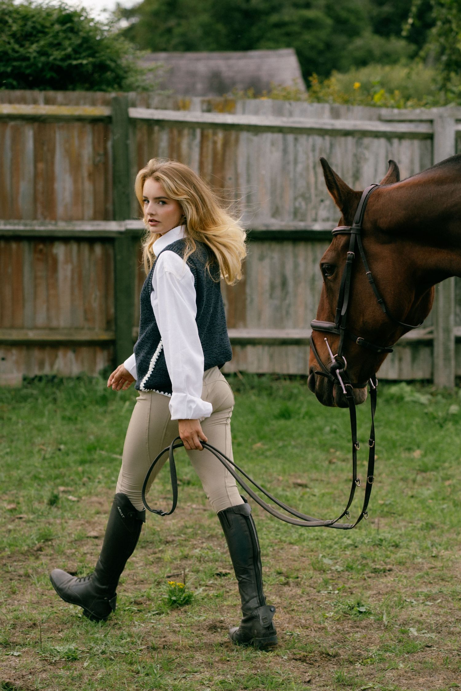 A young woman in equestrian attire leads a brown horse by the reins in a rustic paddock setting.