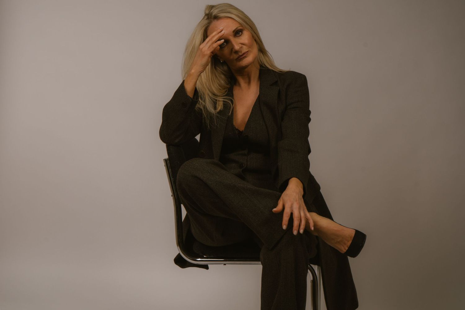 Woman in black suit sitting on chair with hand on head in moody studio portrait.