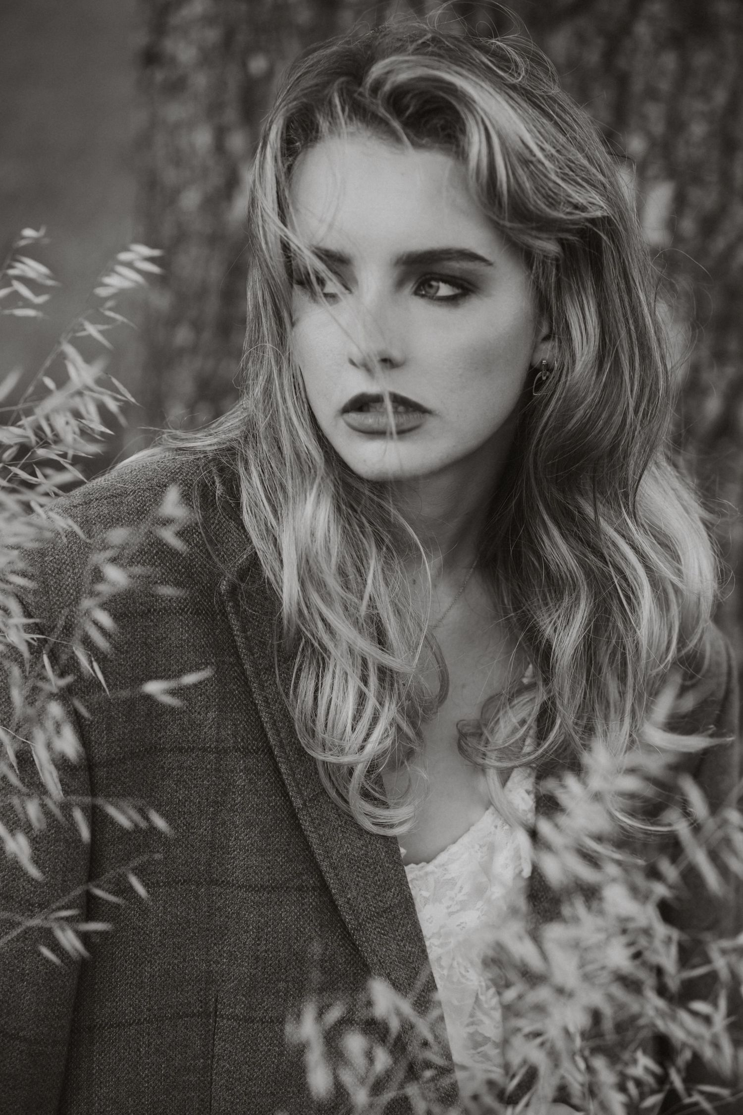 Black and white portrait of a young woman with wavy blonde hair wearing a blazer, surrounded by wild grass outdoors.