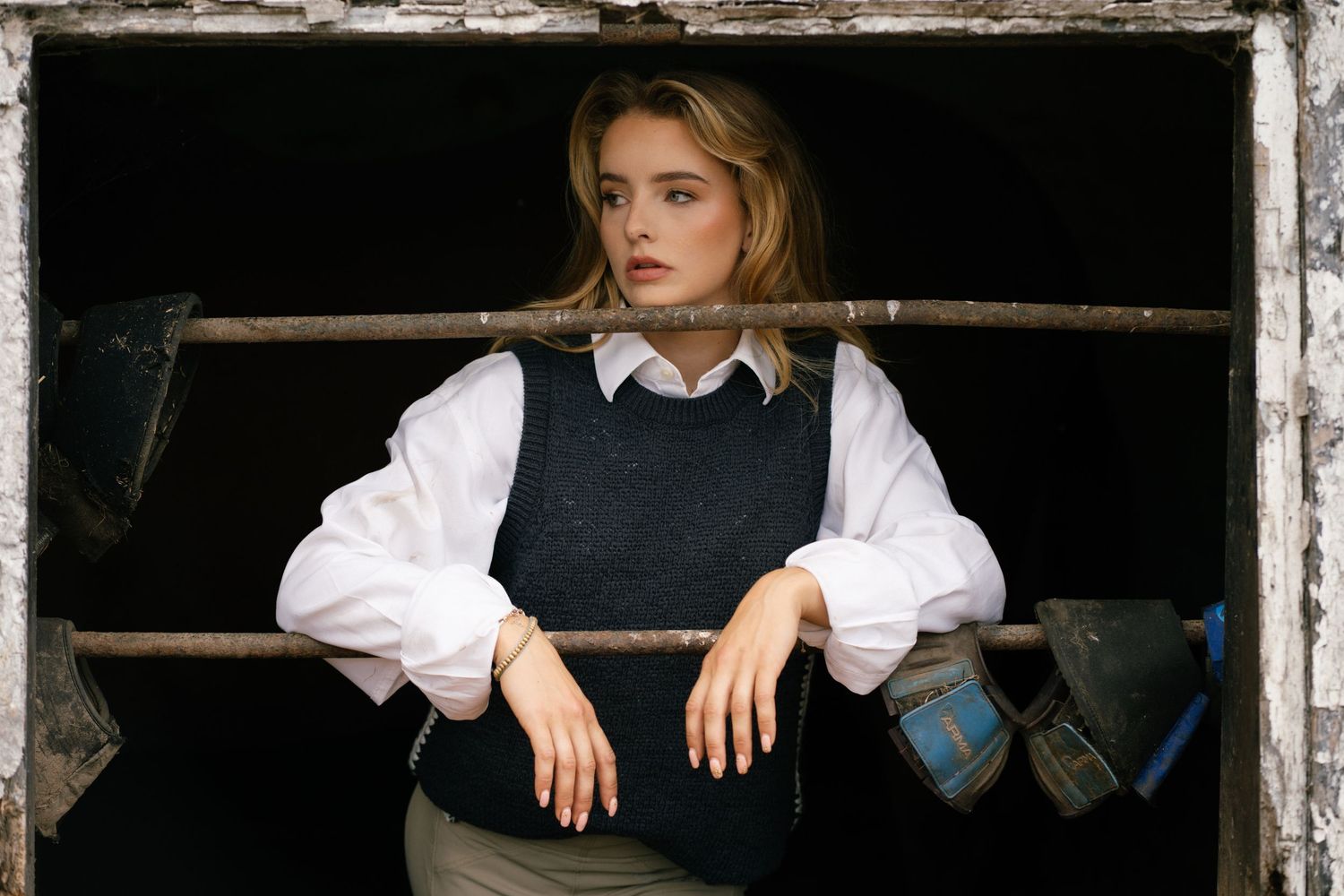 Young woman in white blouse and dark vest leans on wooden frame in rustic setting, looking contemplative.
