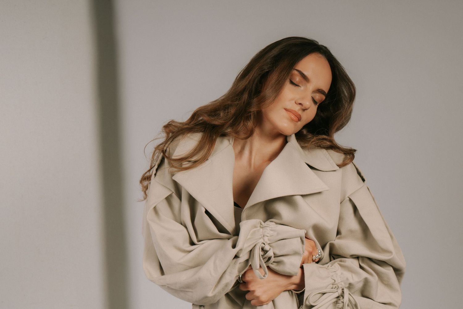 Woman in beige trench coat with eyes closed, flowing brown hair, elegant studio fashion portrait.