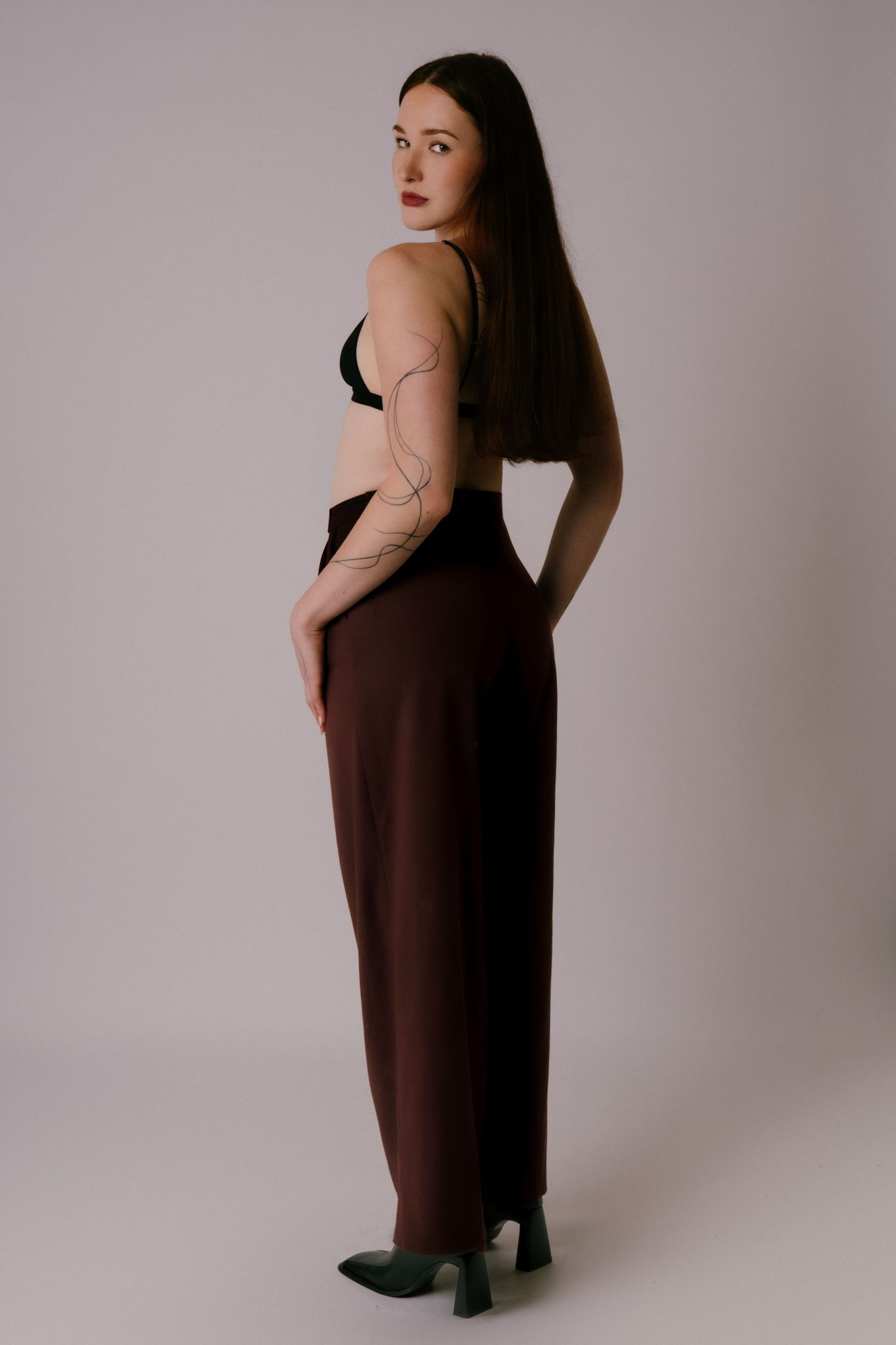 Slender woman in black bra and dark brown wide-leg trousers with body jewelry, posing in profile in studio.