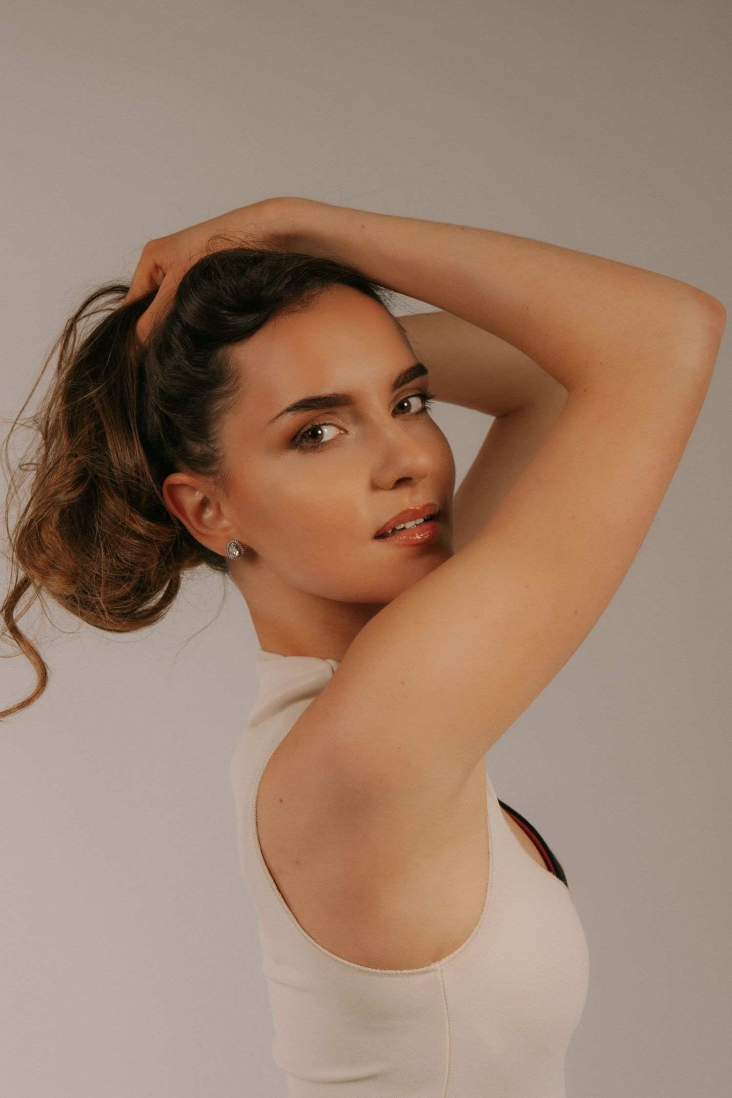 Woman with upswept brown hair in white tank top, arms raised gracefully, clean studio portrait.