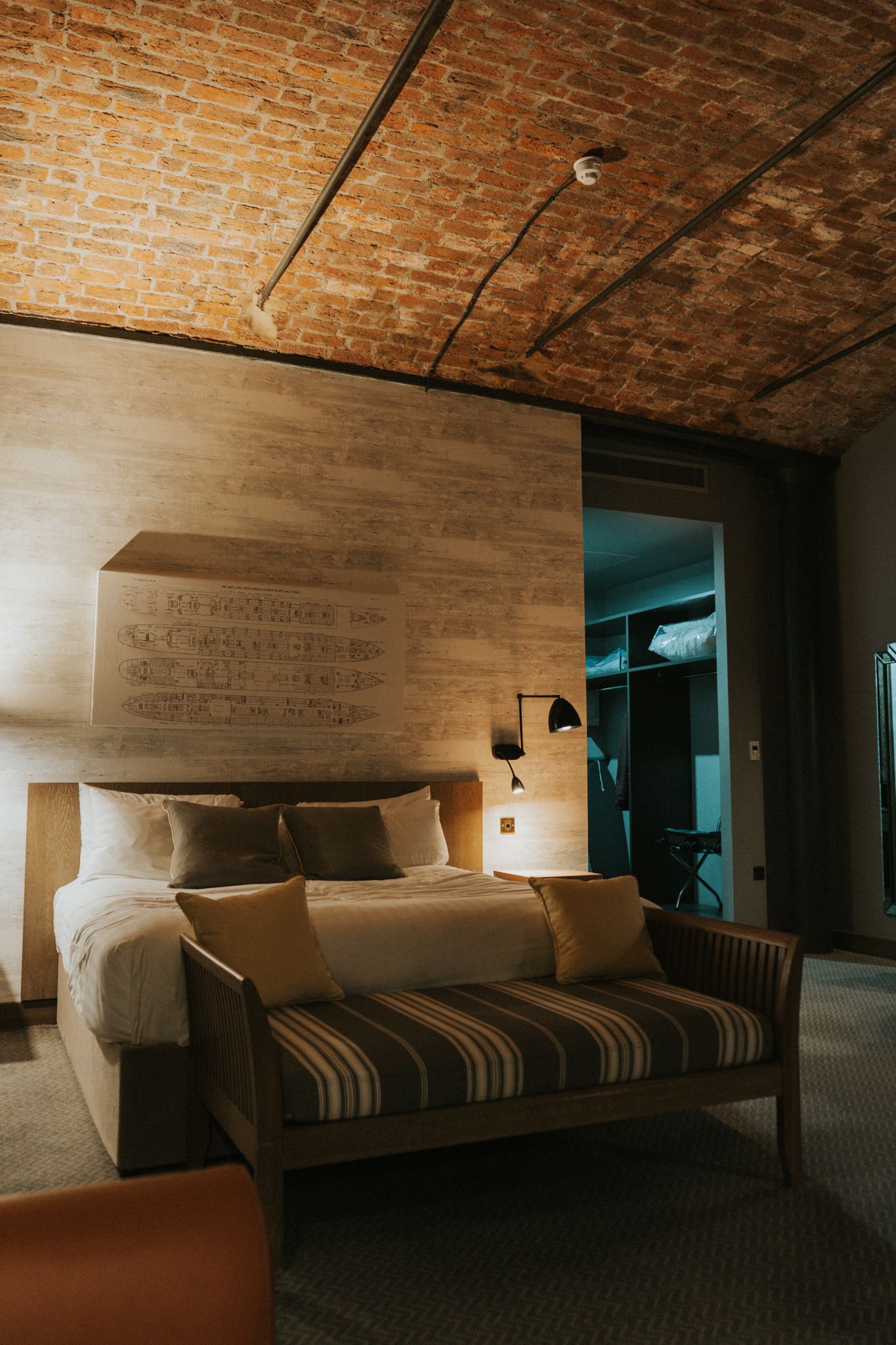 Stylish hotel room with exposed brick ceiling, textured headboard wall, striped bench, and warm lighting.