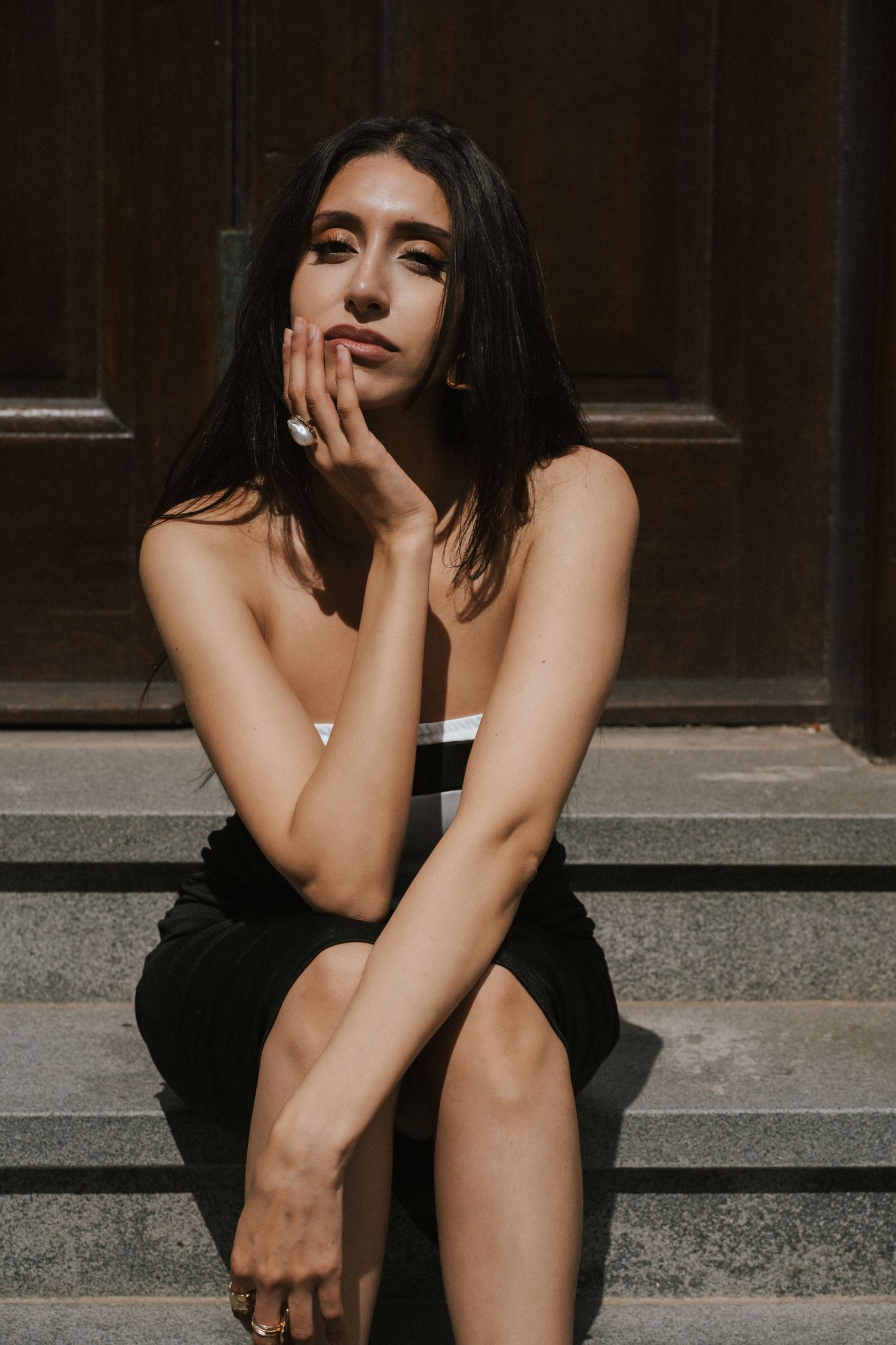 Young woman in black strapless dress sitting on stone steps, posing thoughtfully with hand on face in sunlight.