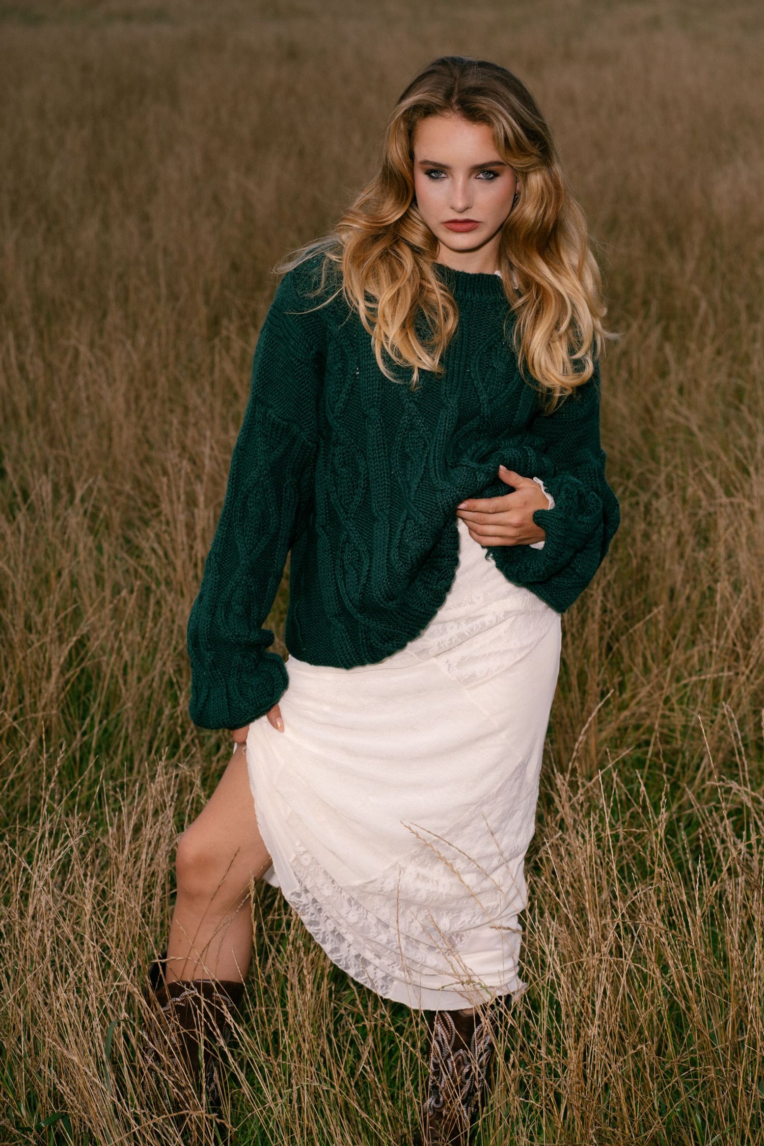 Woman in green cable-knit sweater and white lace midi skirt standing in a golden grass field wearing brown boots.