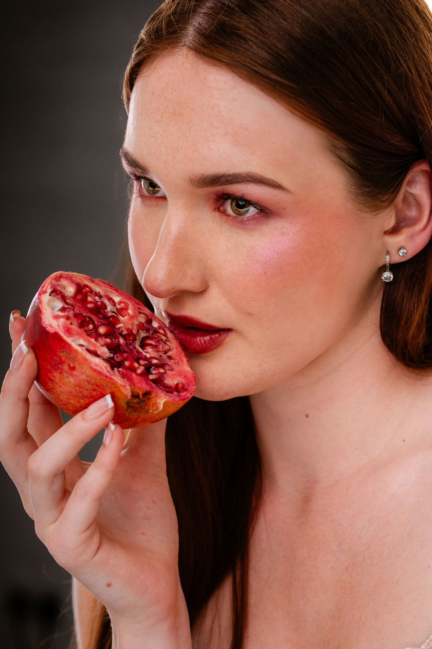 Beautiful woman with dark red lips and freckles holds a ripe pomegranate half against her cheek.