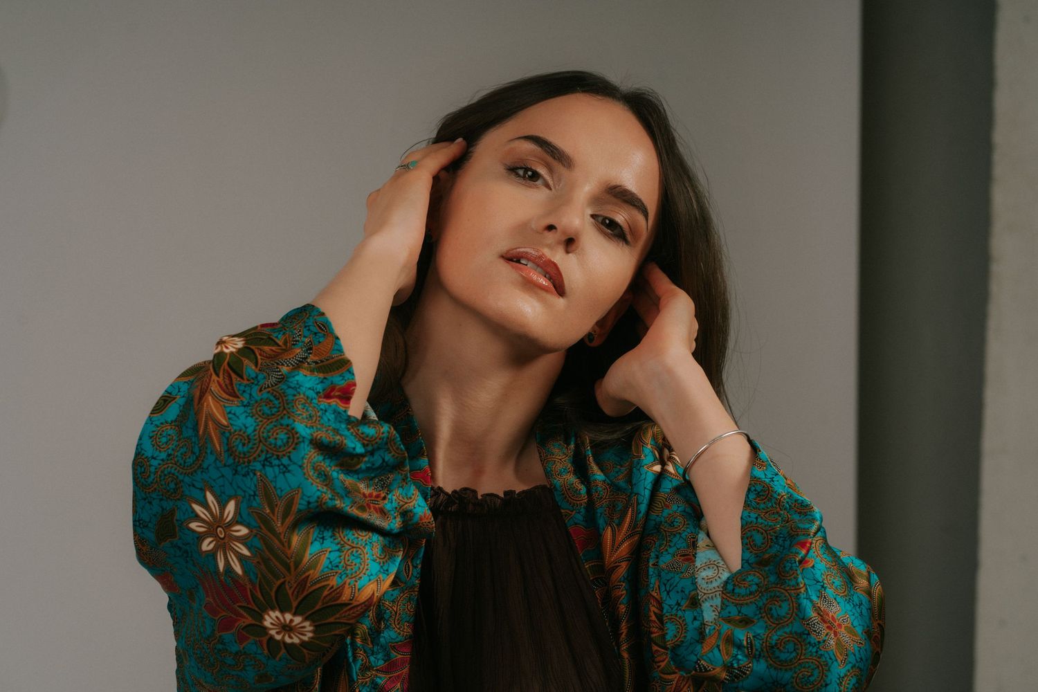 Woman in teal floral jacket with hands framing face, artistic close-up portrait in natural light.