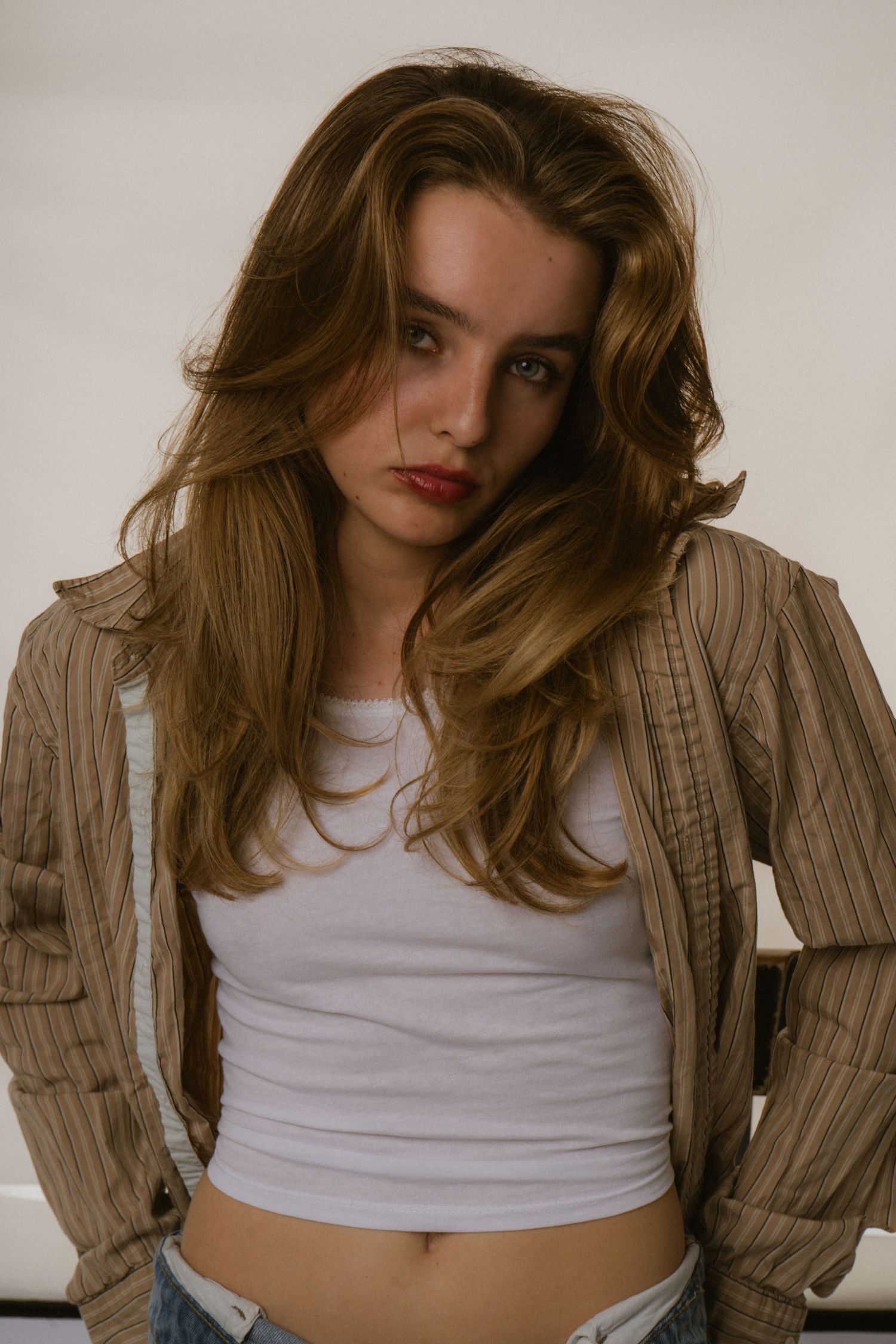 Young woman with long wavy hair in white crop top and open striped jacket poses against white background.