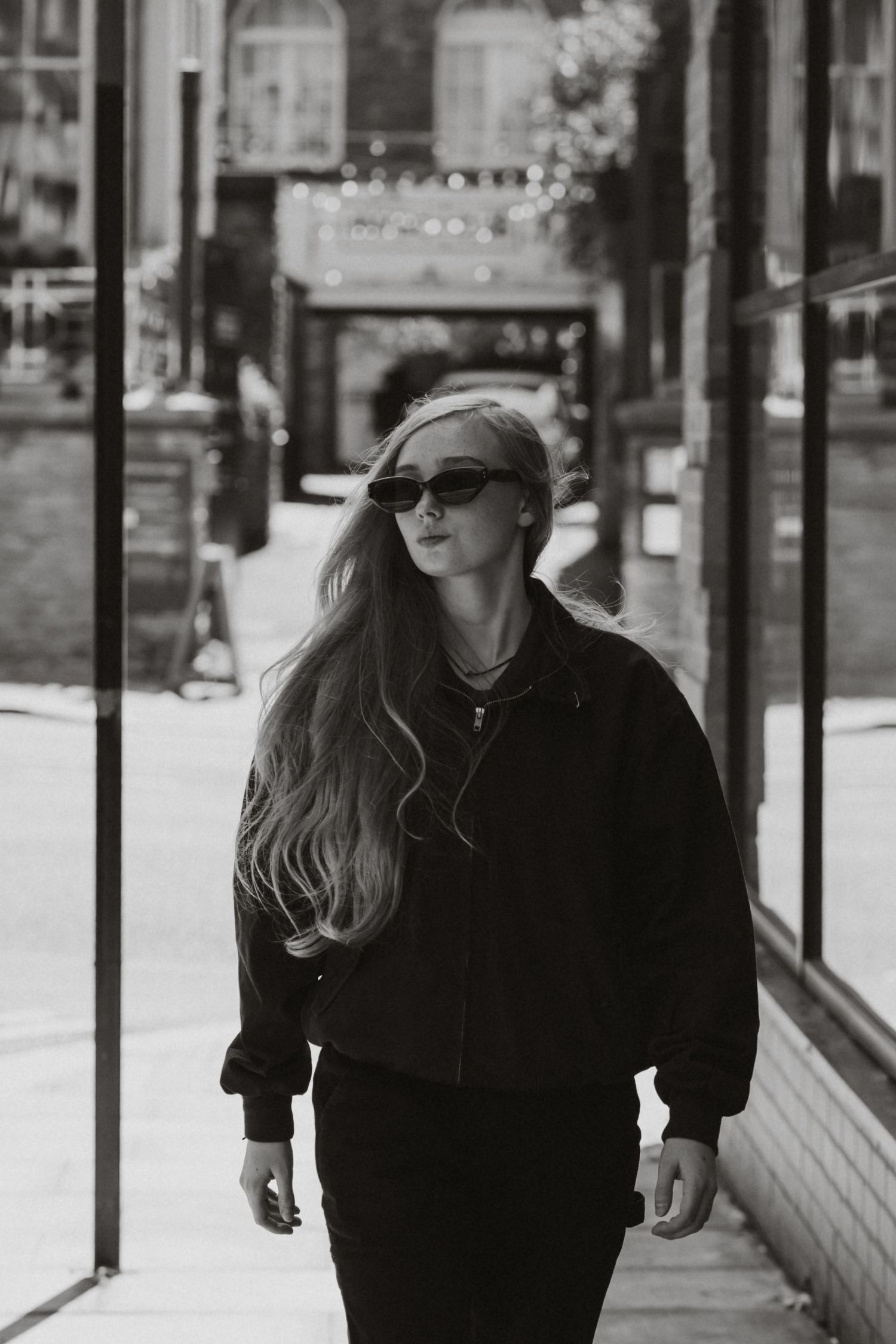 Black and white photo of a young woman with long hair wearing sunglasses and a bomber jacket walking on a city sidewalk.