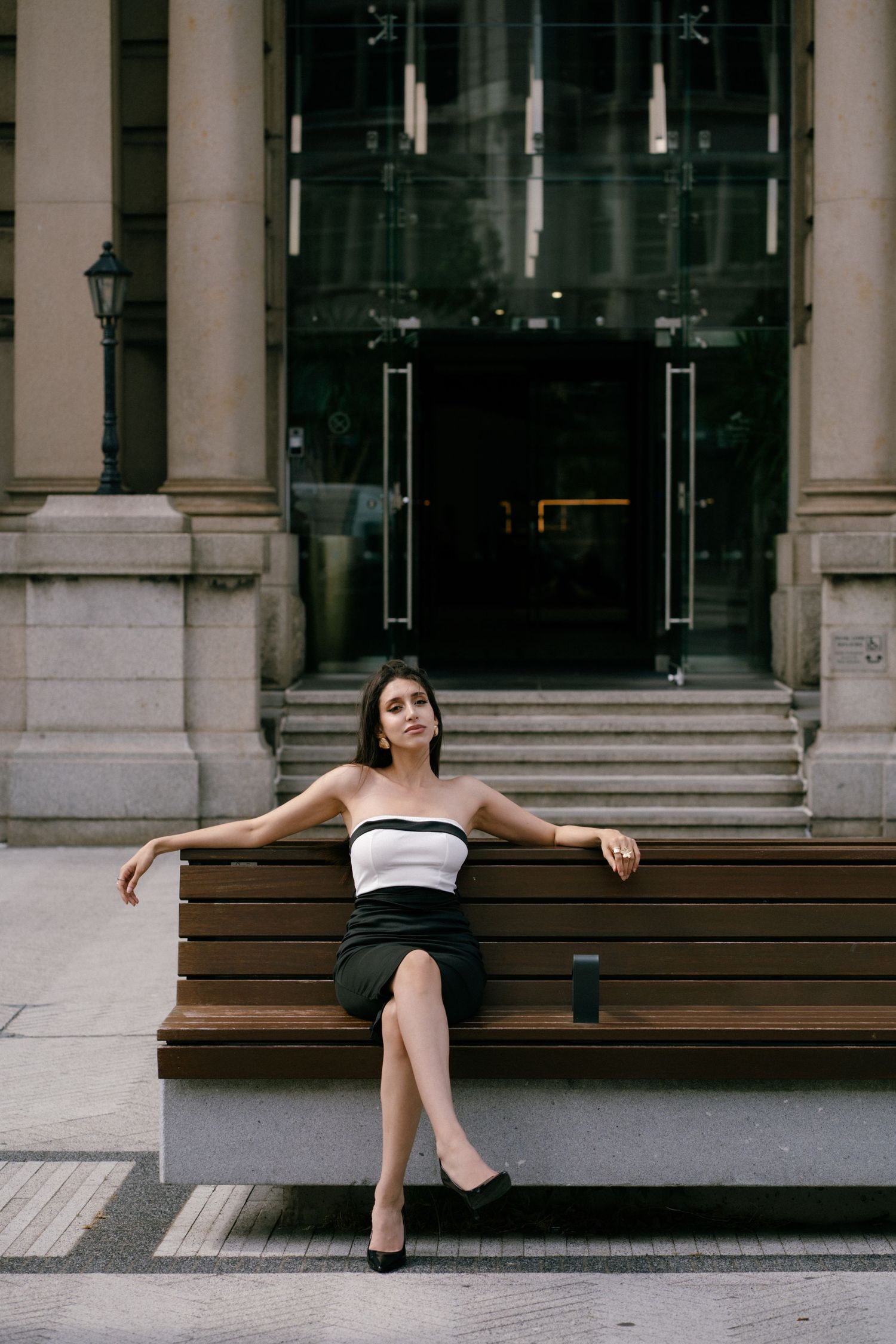 Elegant woman in black skirt and white strapless top sitting on wooden bench outside classic building.