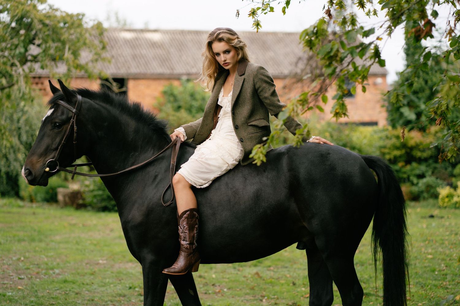 A blonde woman in a white dress, blazer, and cowboy boots sits elegantly on a black horse near a rustic barn.
