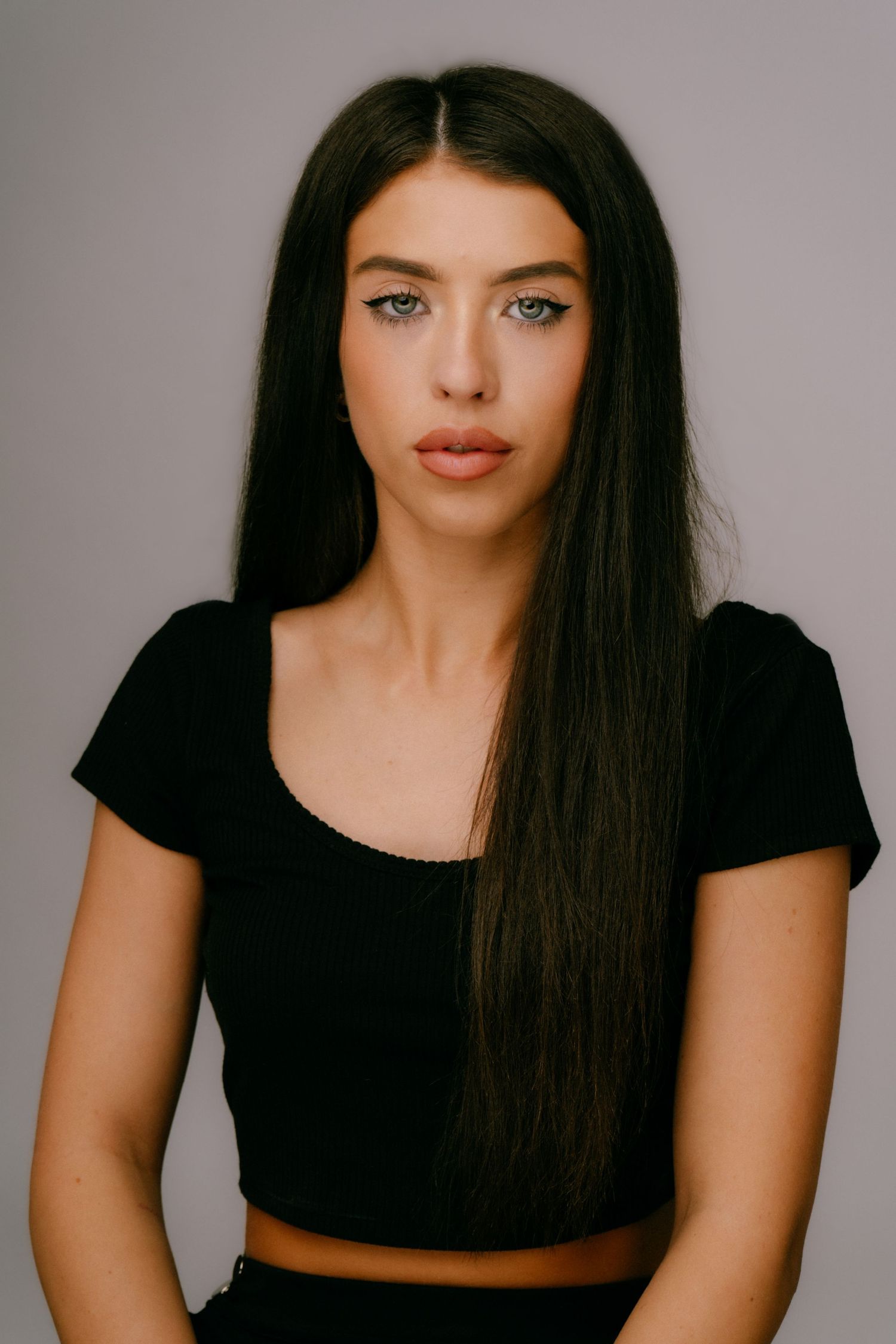 Young woman with long dark hair and blue eyes wearing a black crop top poses against a gray background.