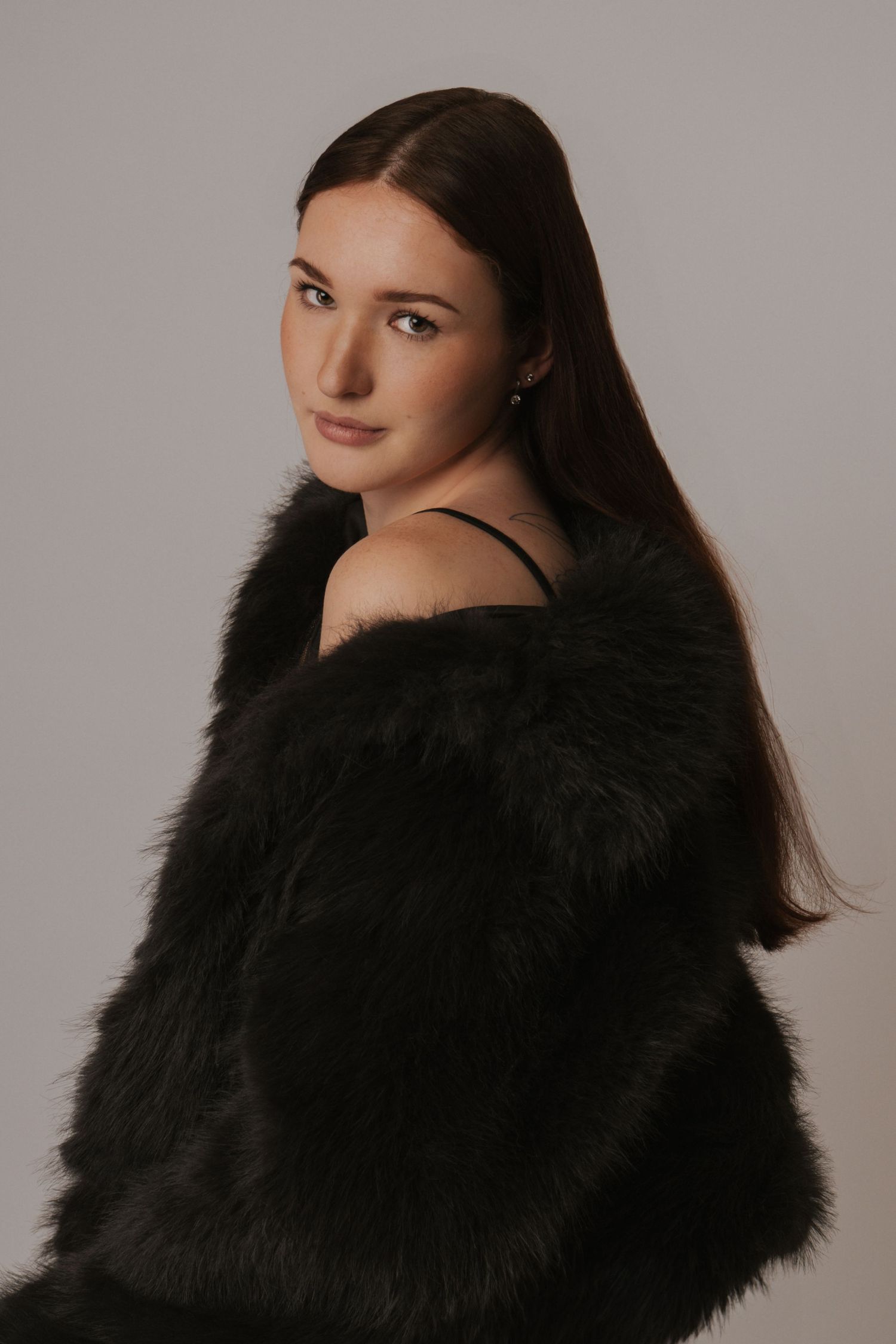 Young brunette woman wearing a black faux fur coat, posing over shoulder against a light gray studio background.