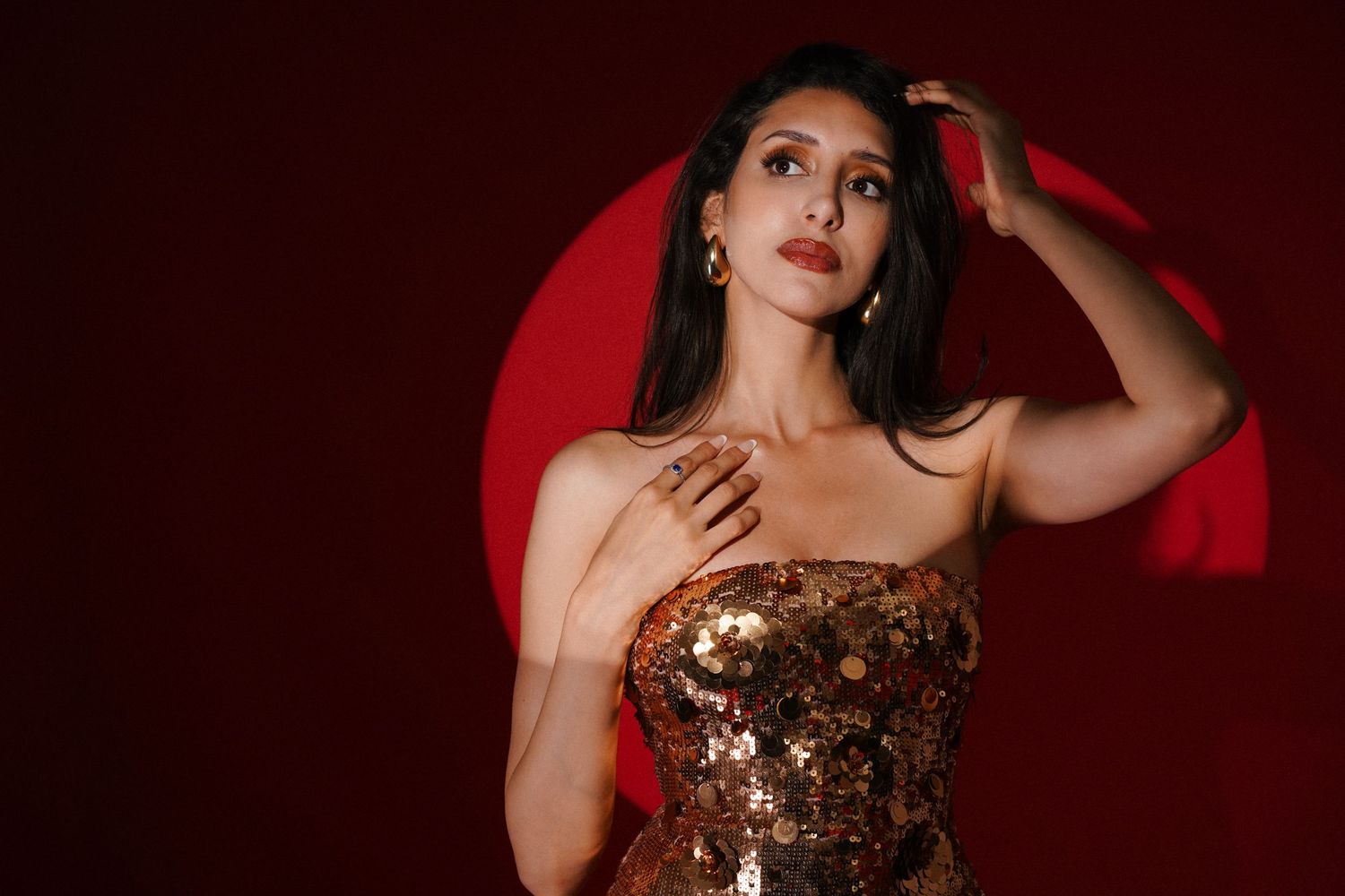 Woman in gold sequin dress poses against red background with circular light, hand on chest and hair.