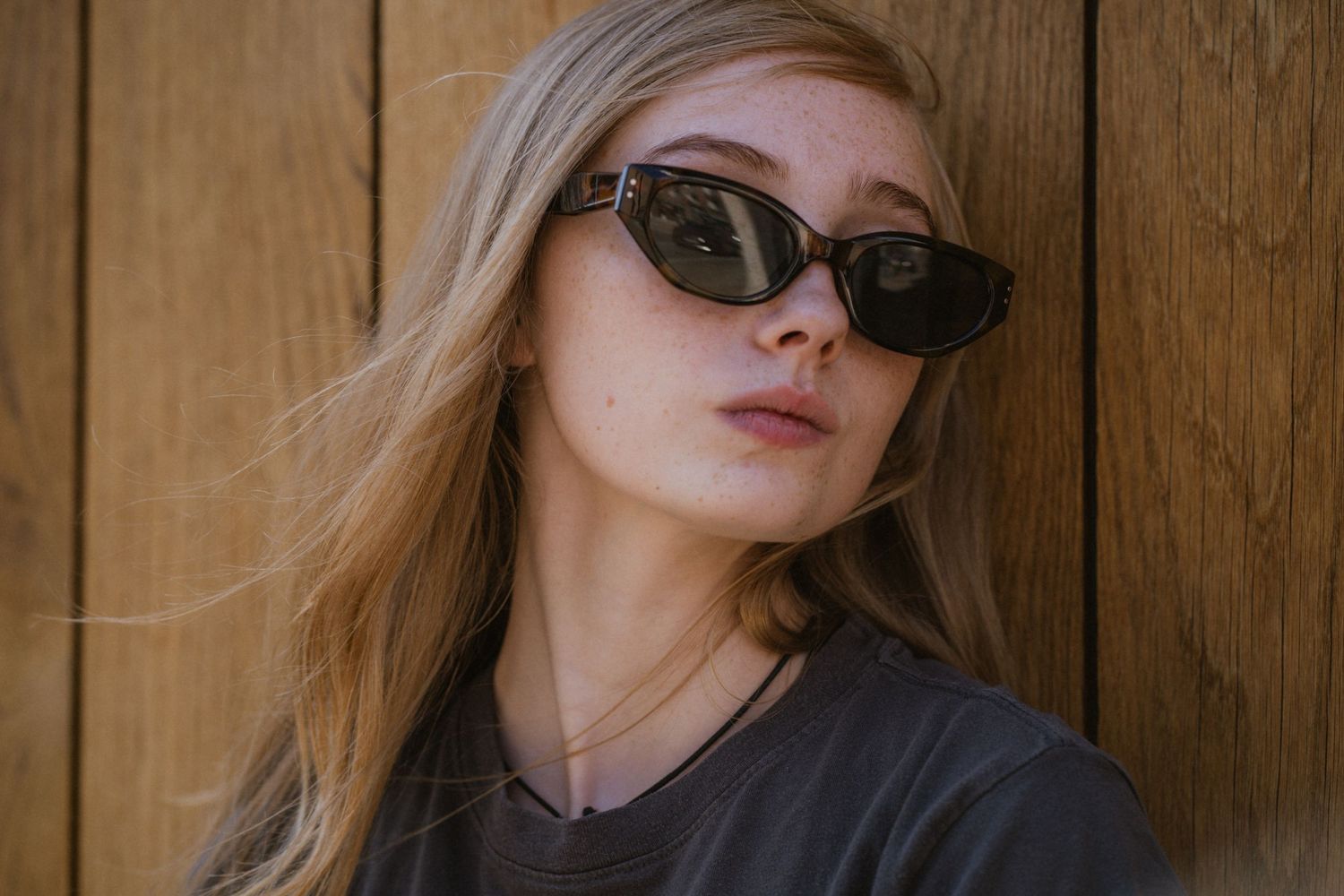 Young blonde woman wearing dark oval sunglasses leaning against a wooden wall in a gray t-shirt.