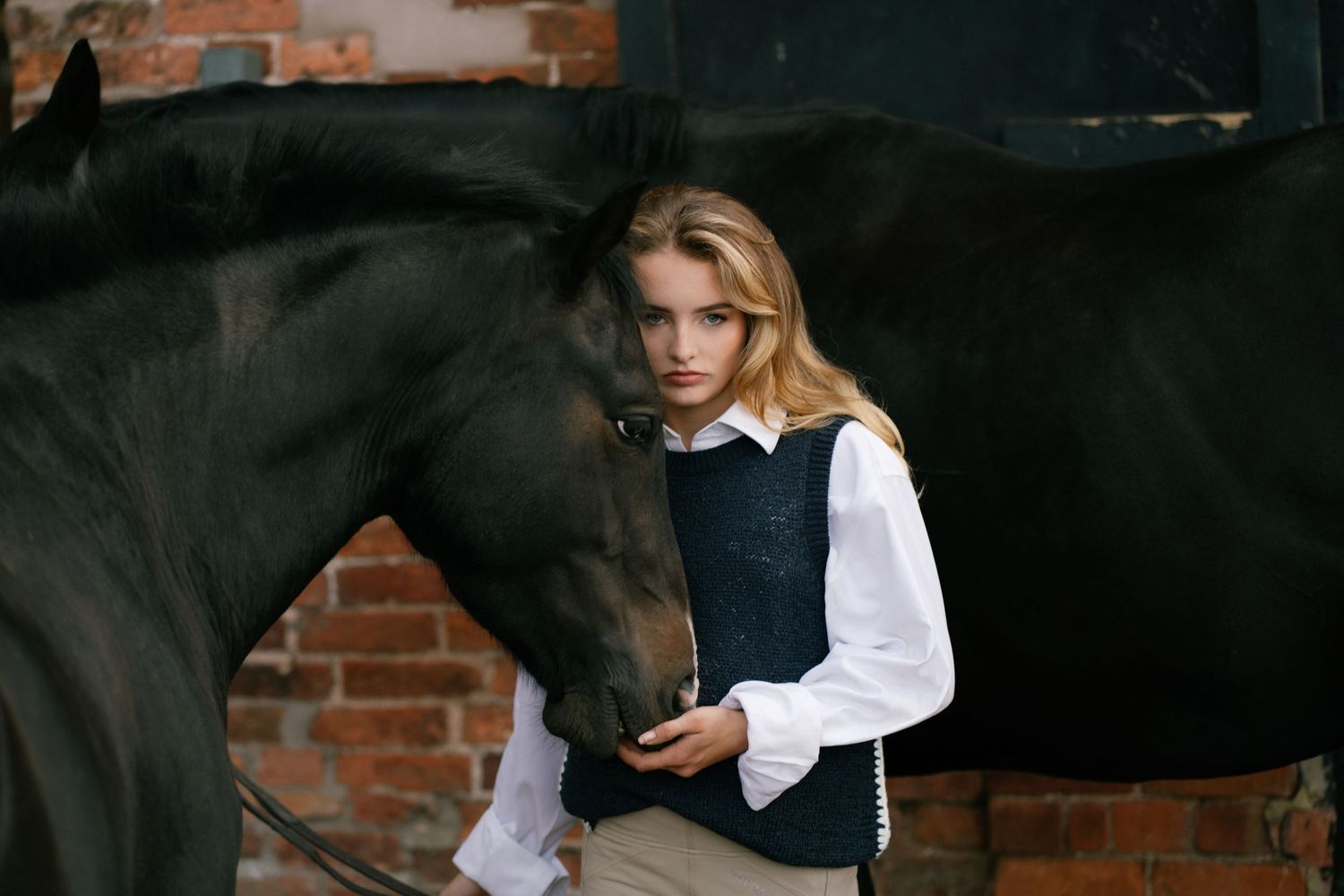 Young blonde girl in equestrian attire standing beside a dark horse near a brick wall.
