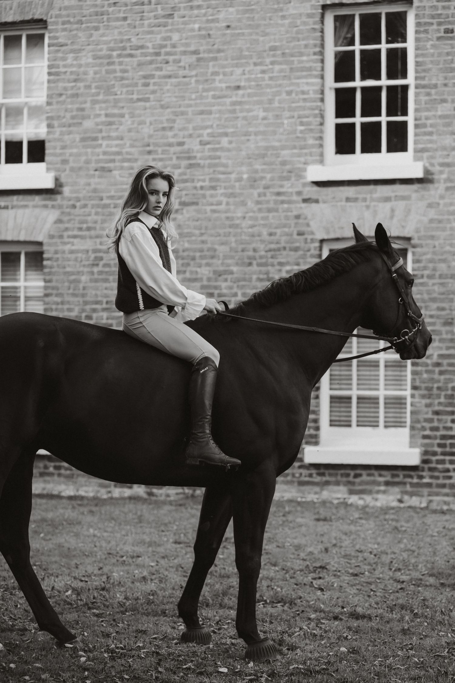 Black and white photo of a young woman in equestrian attire riding a dark horse beside a brick building.