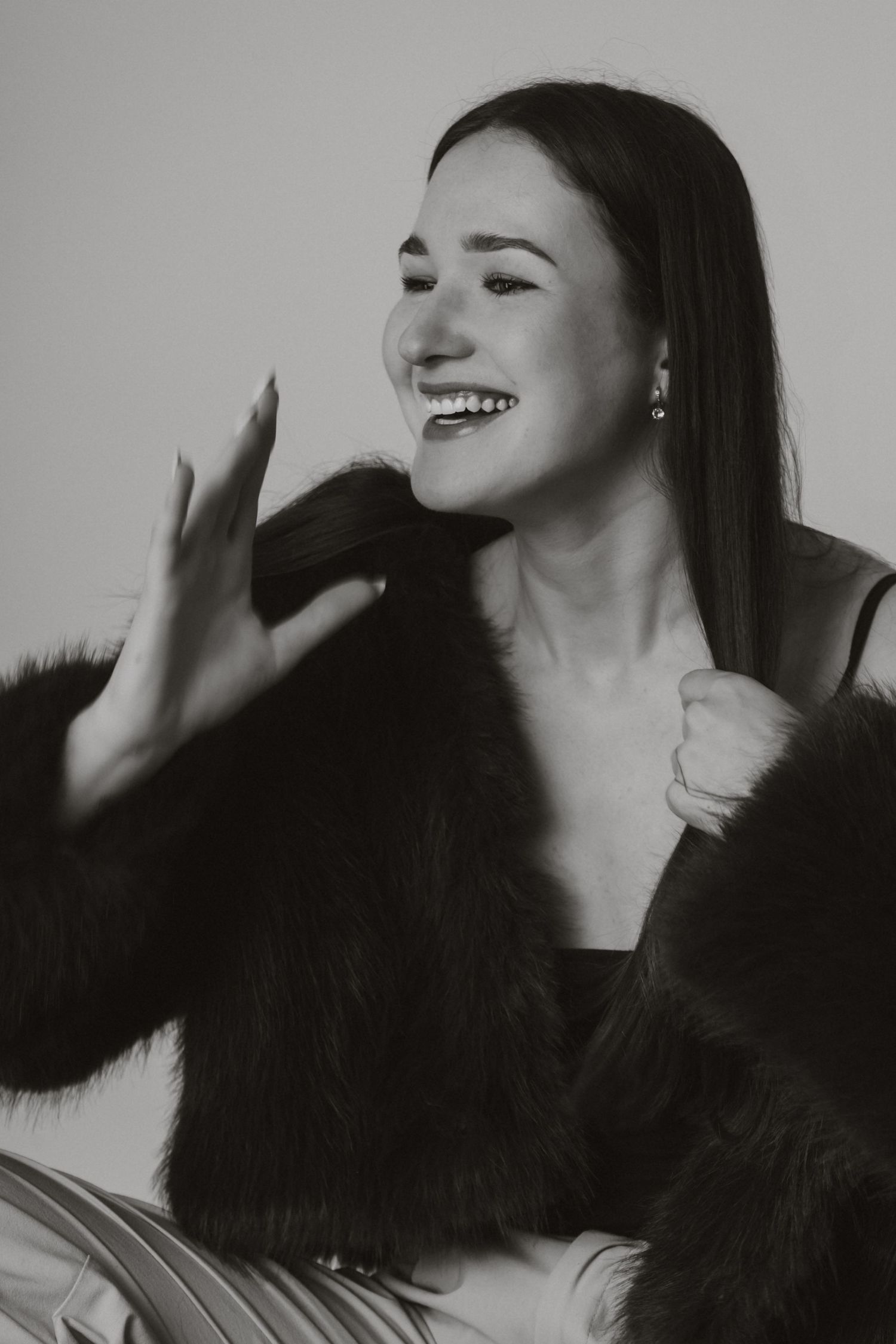 Black and white photo of laughing young woman in black faux fur coat touching her hair in a studio setting.