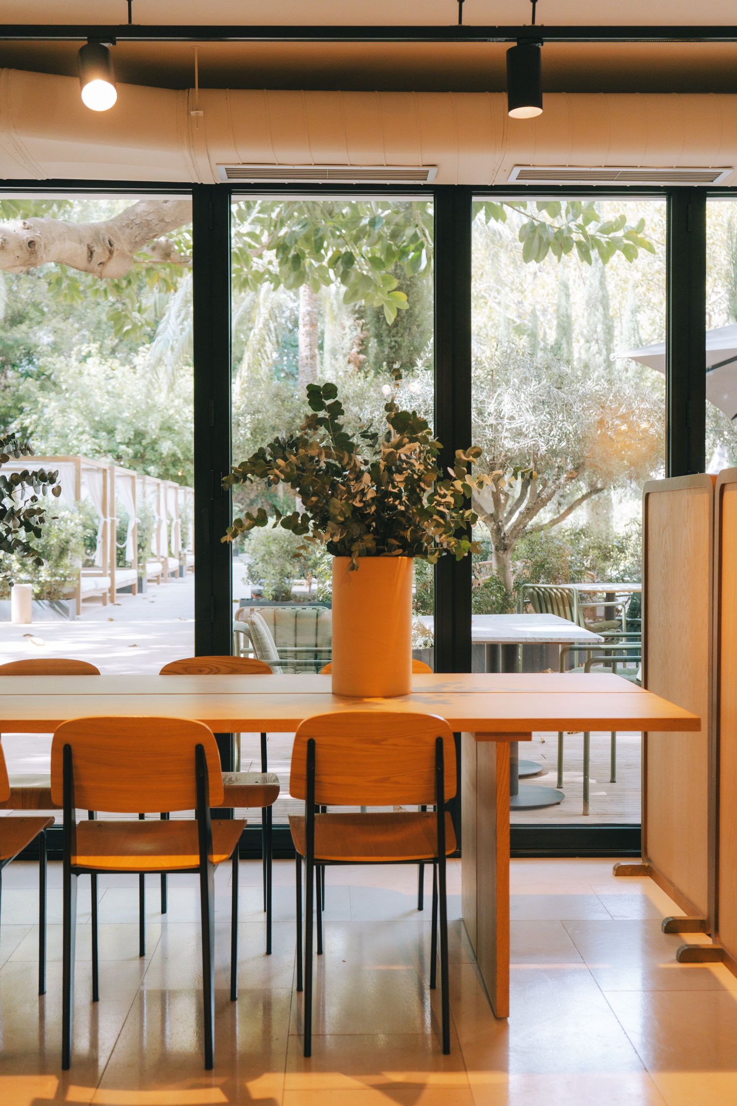 Modern cafe dining area with wooden chairs, long table, orange vase with greenery, and large windows overlooking lush trees.