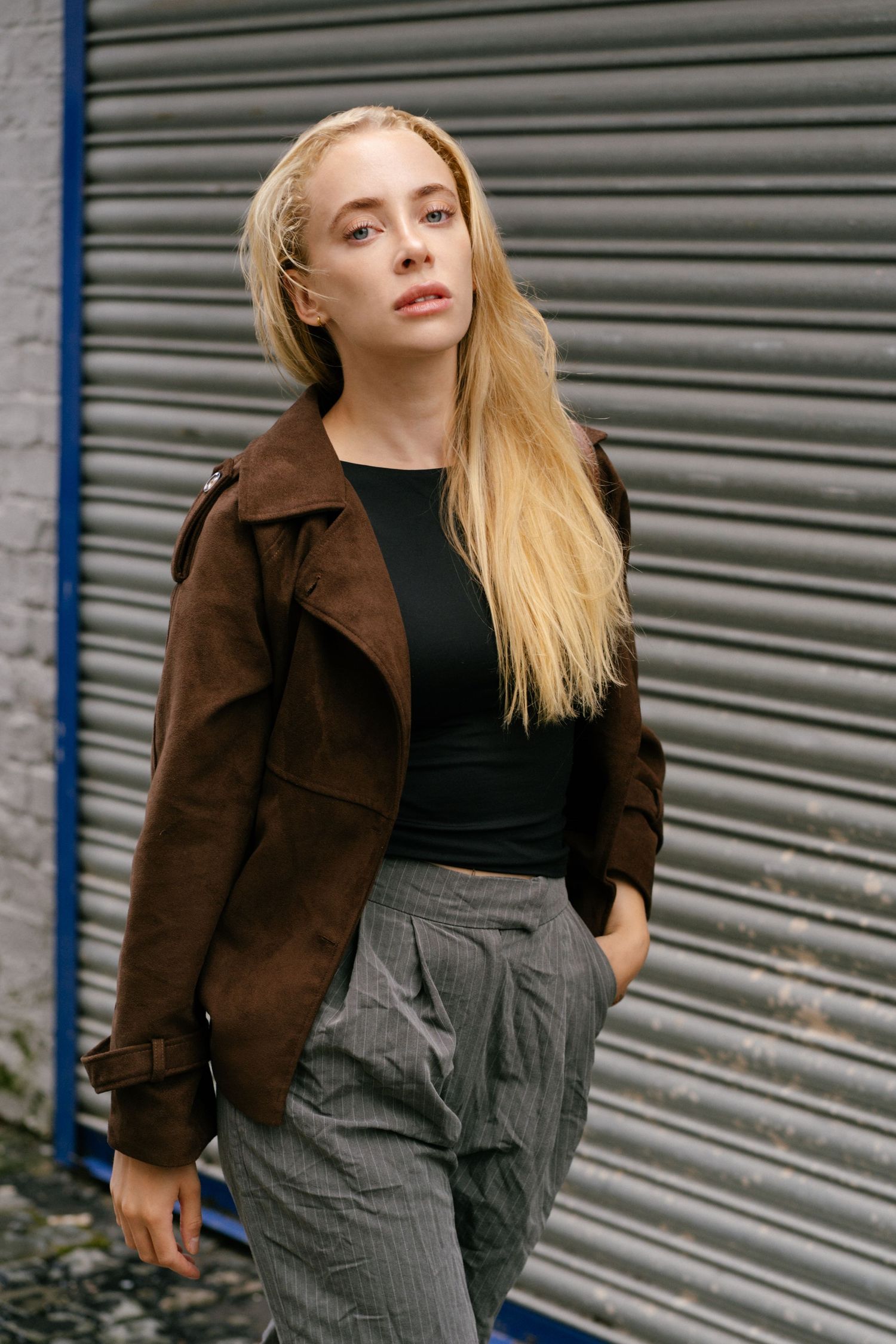 Blonde woman in brown suede jacket, black top, and gray pinstripe trousers posing against a metal shutter.