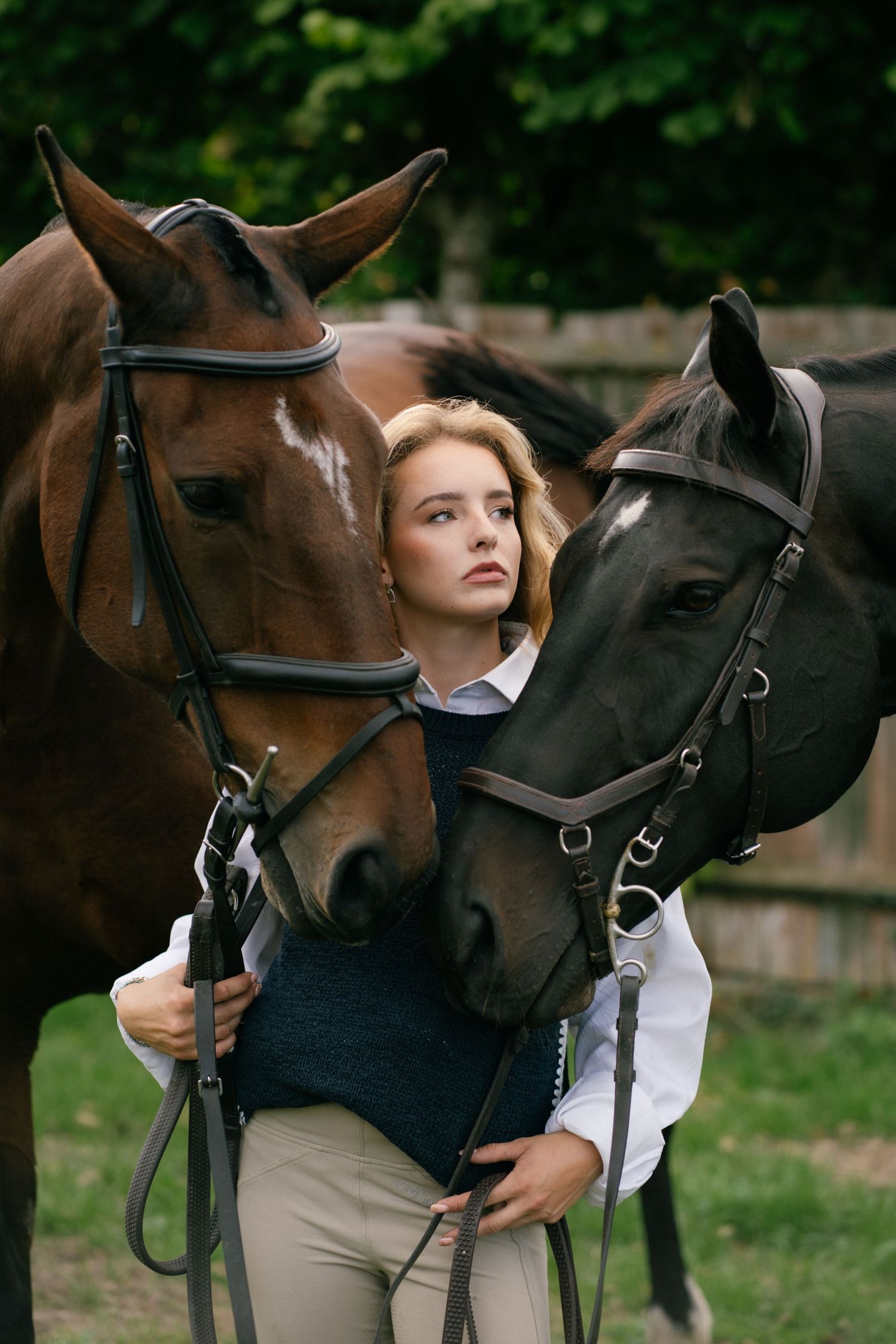 Young blonde woman standing between two horses, a brown and a black, holding their reins outdoors.