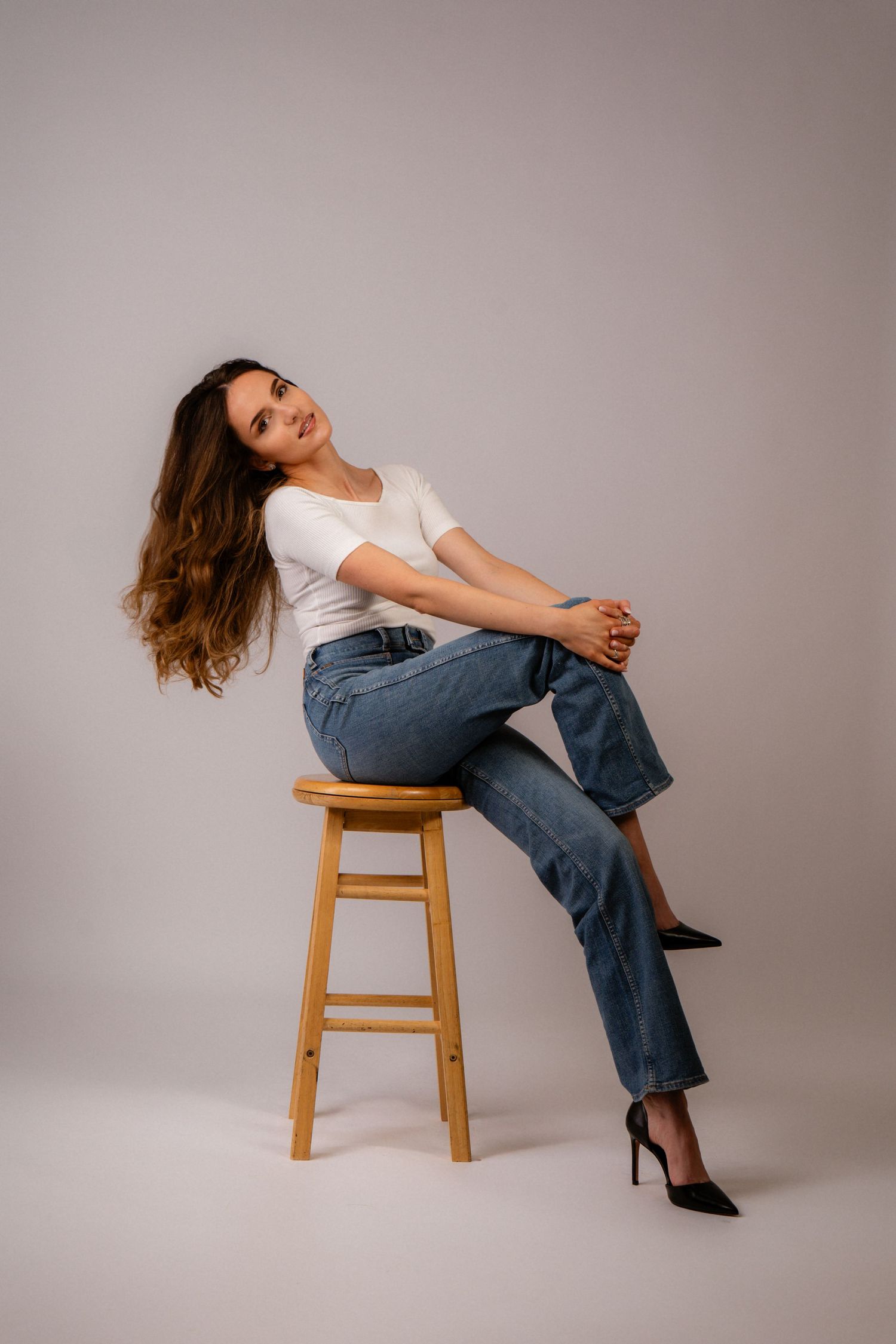 Woman in white t-shirt and blue jeans sitting on wooden stool with flowing hair, studio fashion photo.
