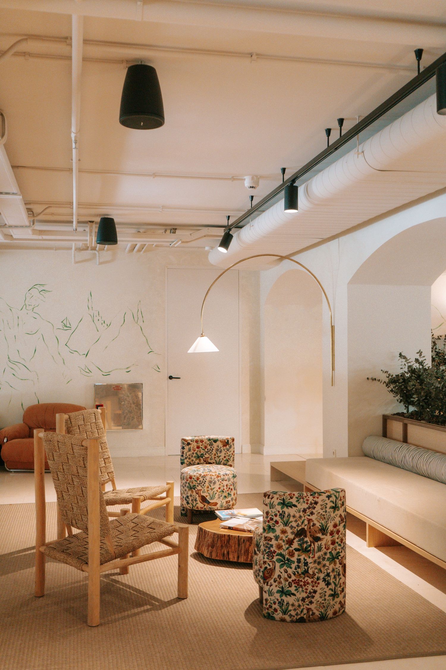 Stylish hotel lounge with floral armchairs, woven chairs, arched walls, arc floor lamp, and botanical wall mural.
