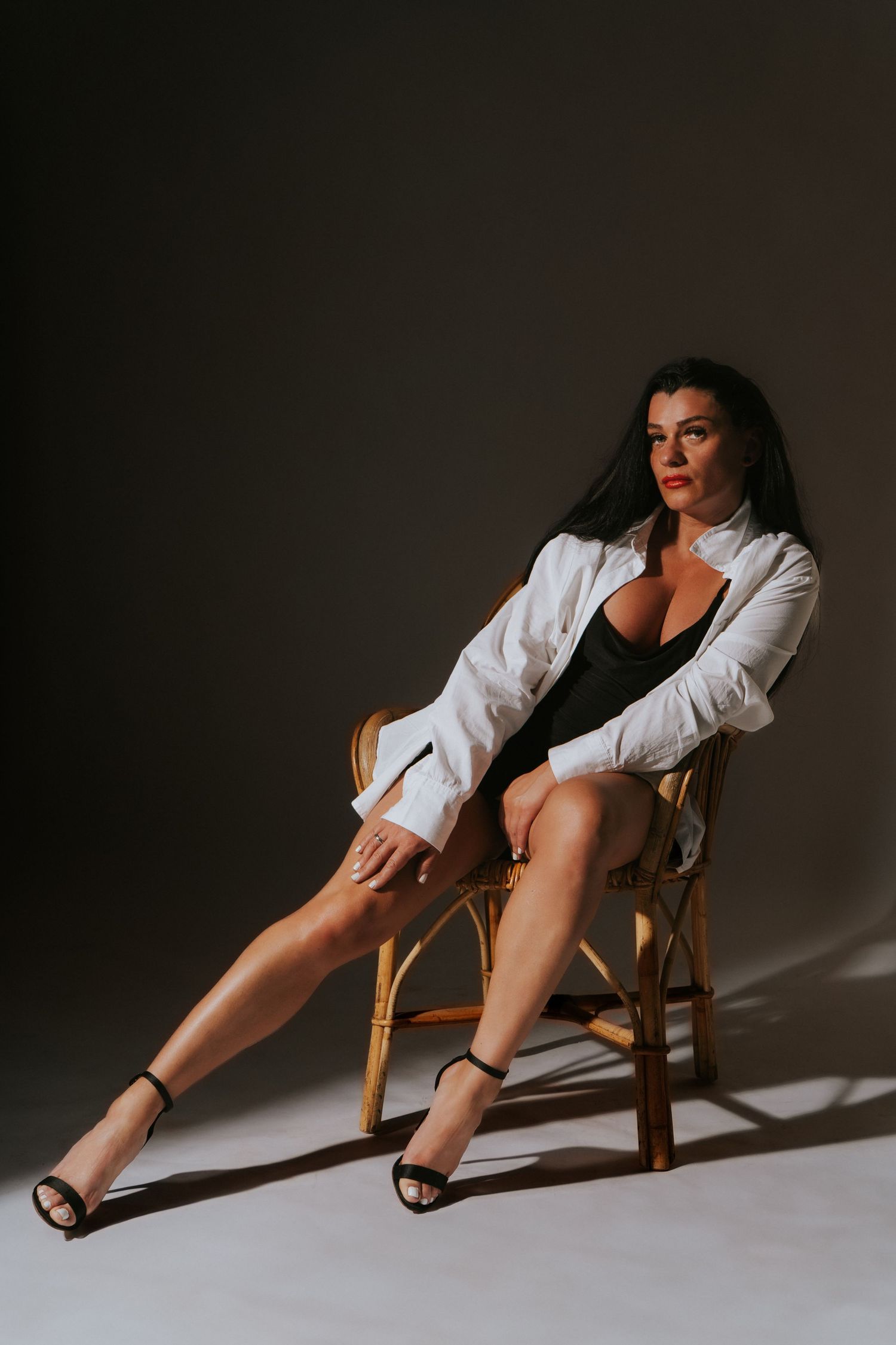 Woman in white blazer and black bodysuit seated in wooden chair in moody studio setting.