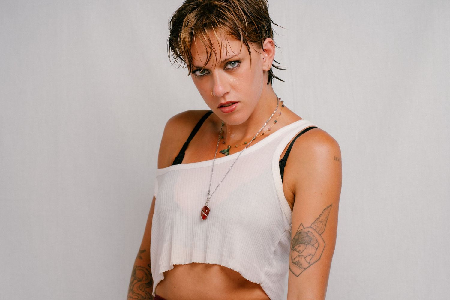 Young woman with wet hair, tattoos, and necklaces poses in a white crop top against a light background.