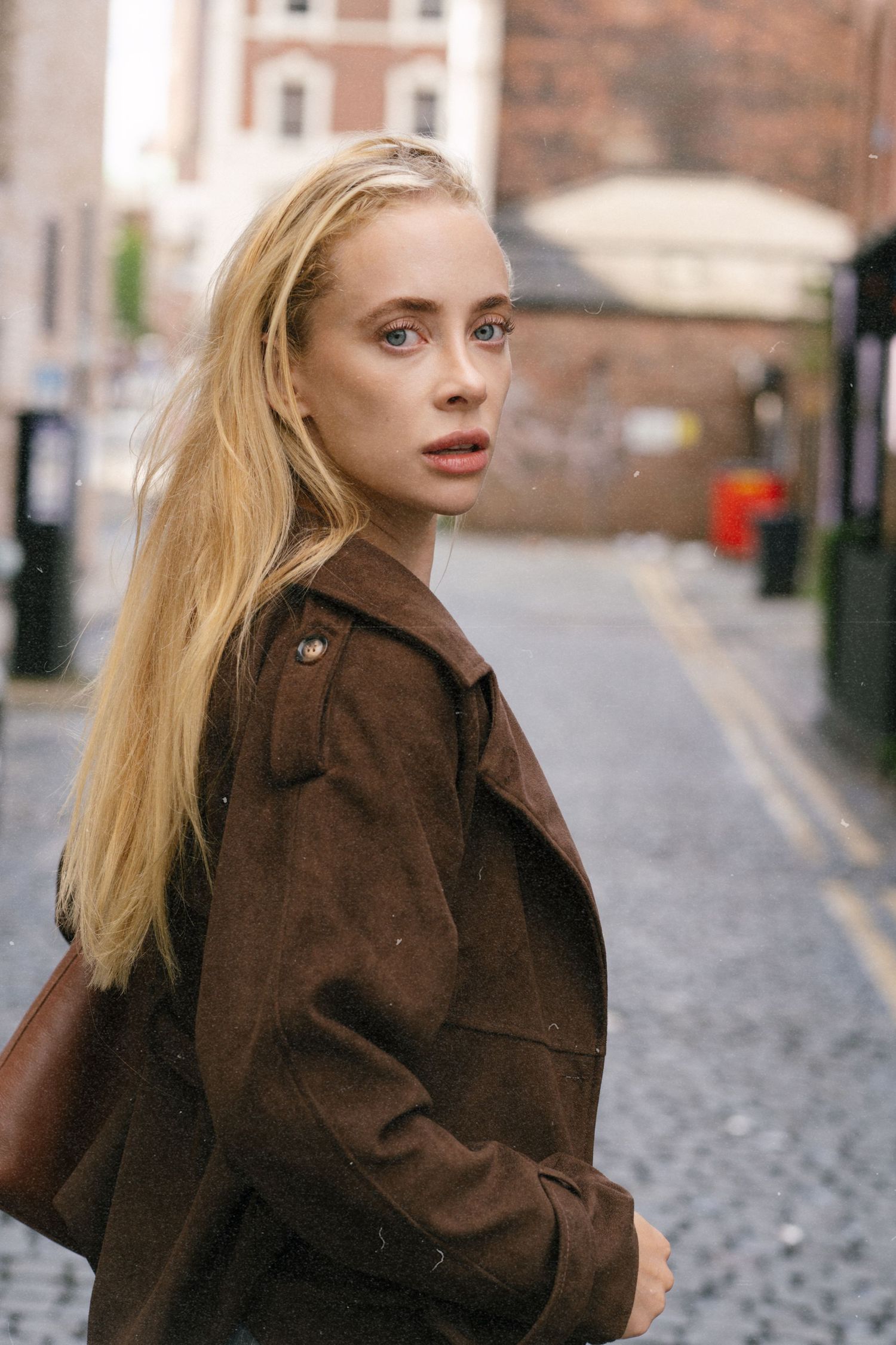 Blonde woman in brown suede jacket looking over shoulder on cobblestone city street.