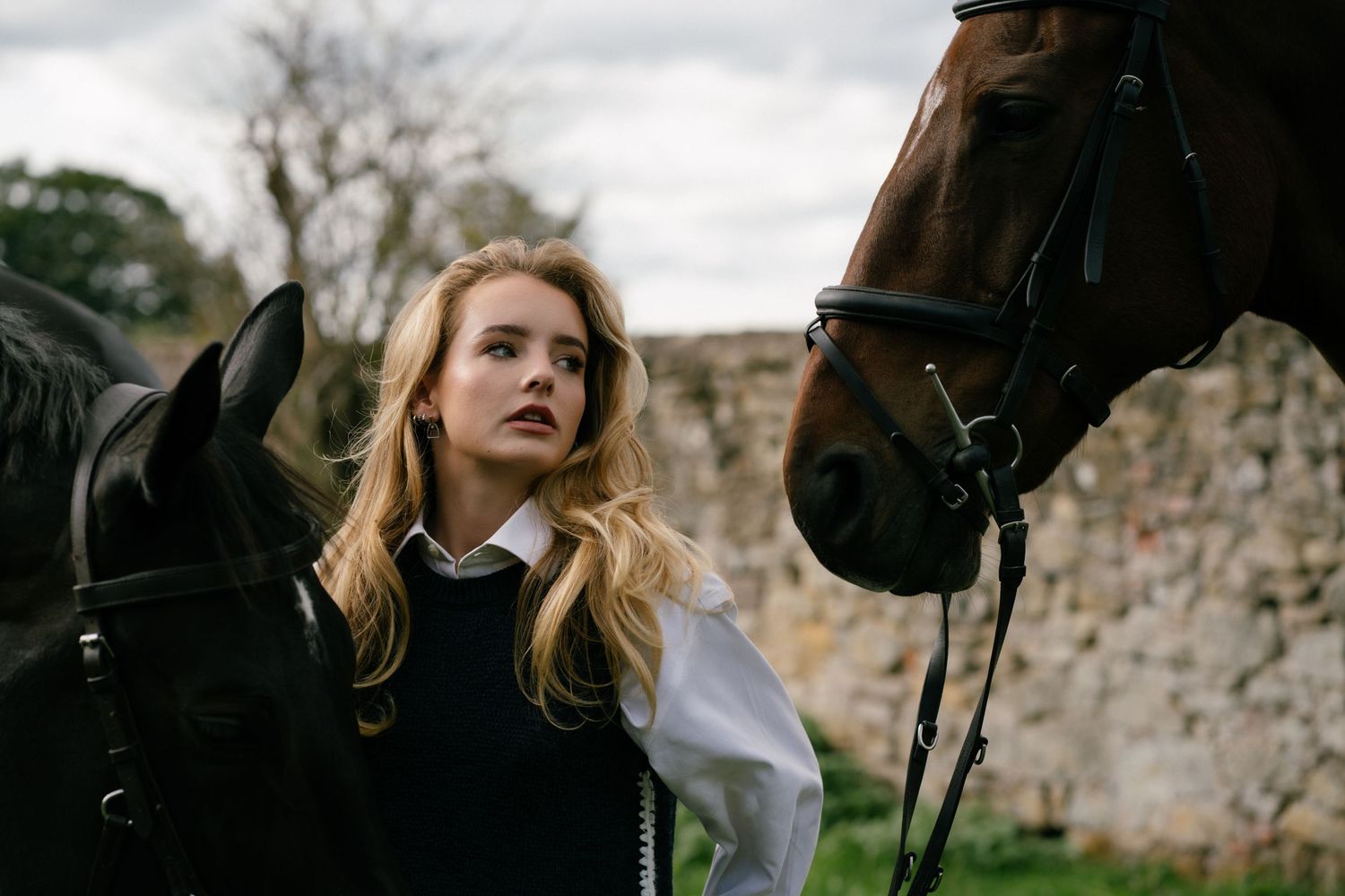 Young blonde woman in equestrian attire standing between two dark horses near a stone wall outdoors.