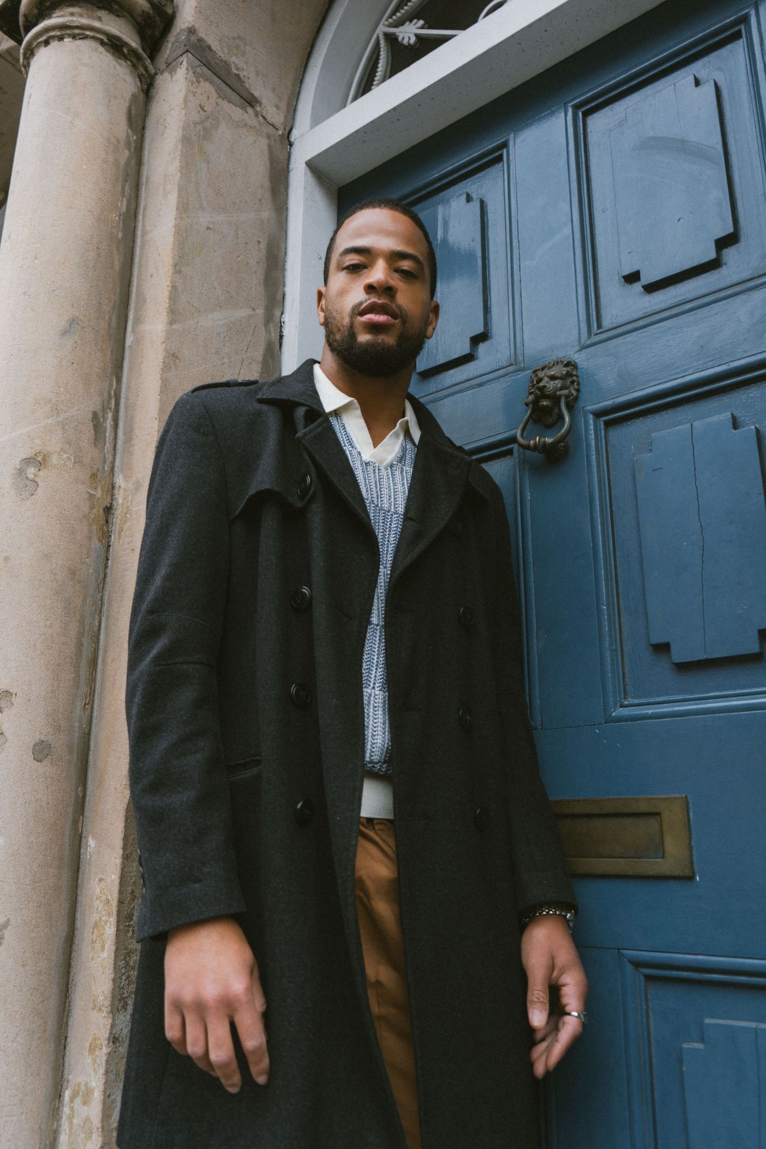 Fashionable man in long black coat standing before blue door with lion knocker.