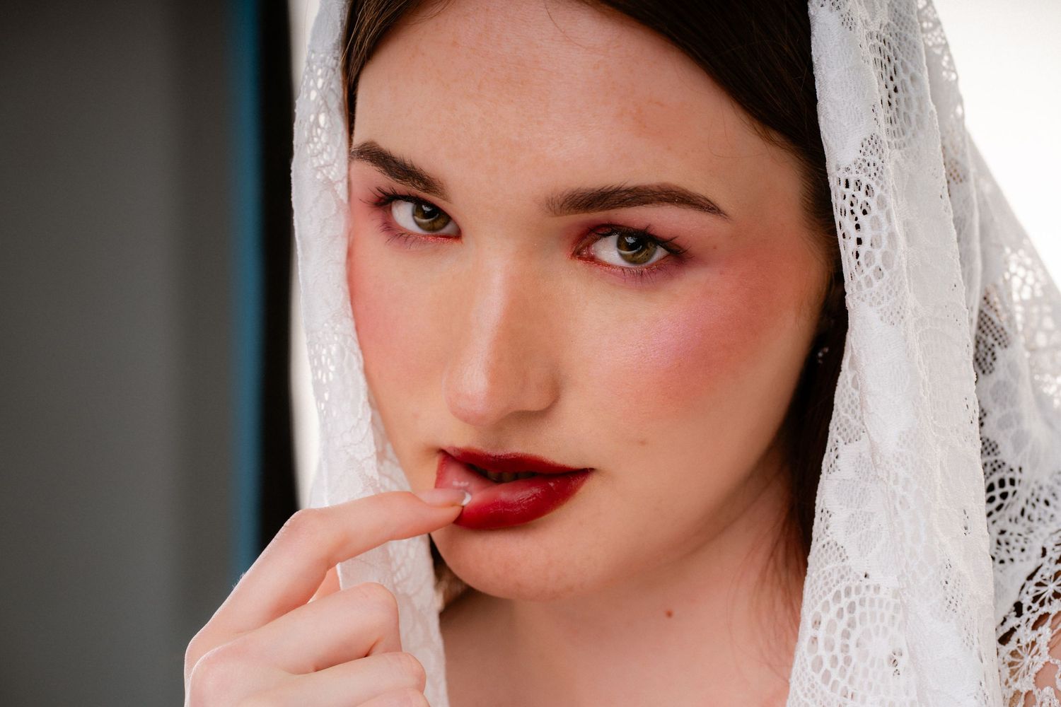 Close-up of a woman with bold red lips and rosy blush wearing a white lace veil, touching her lip.