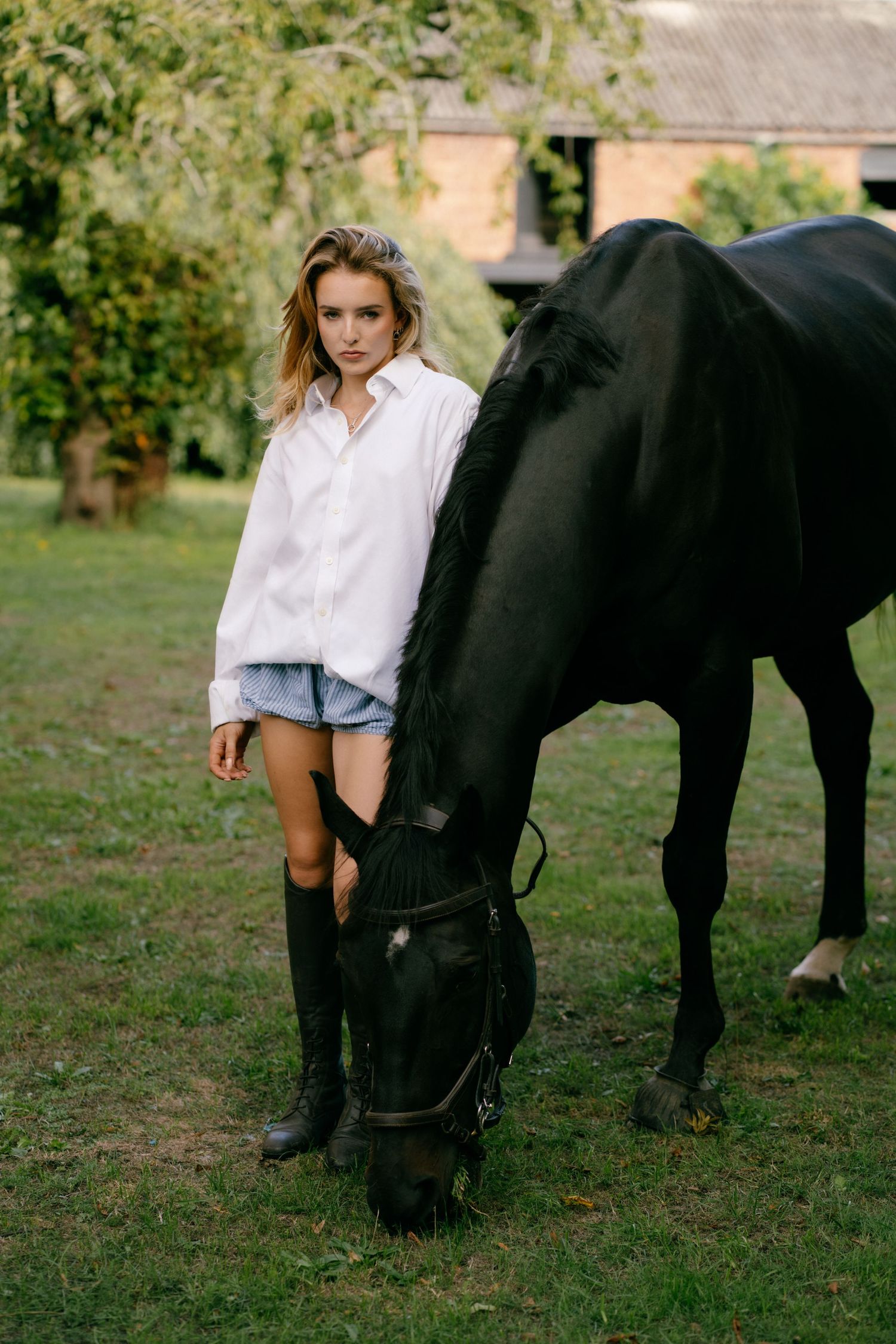 Young woman in white shirt and denim shorts stands beside a grazing black horse on a lush green farm.