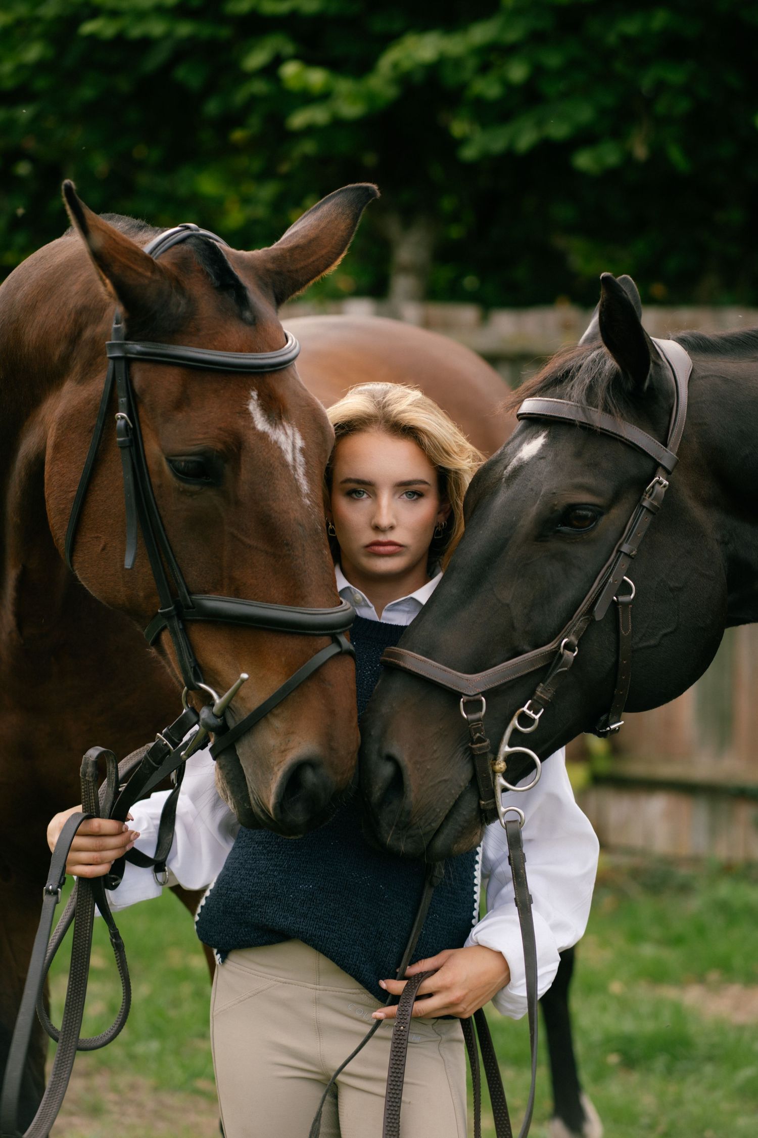 Young blonde woman in equestrian attire standing between two brown horses wearing bridles outdoors.