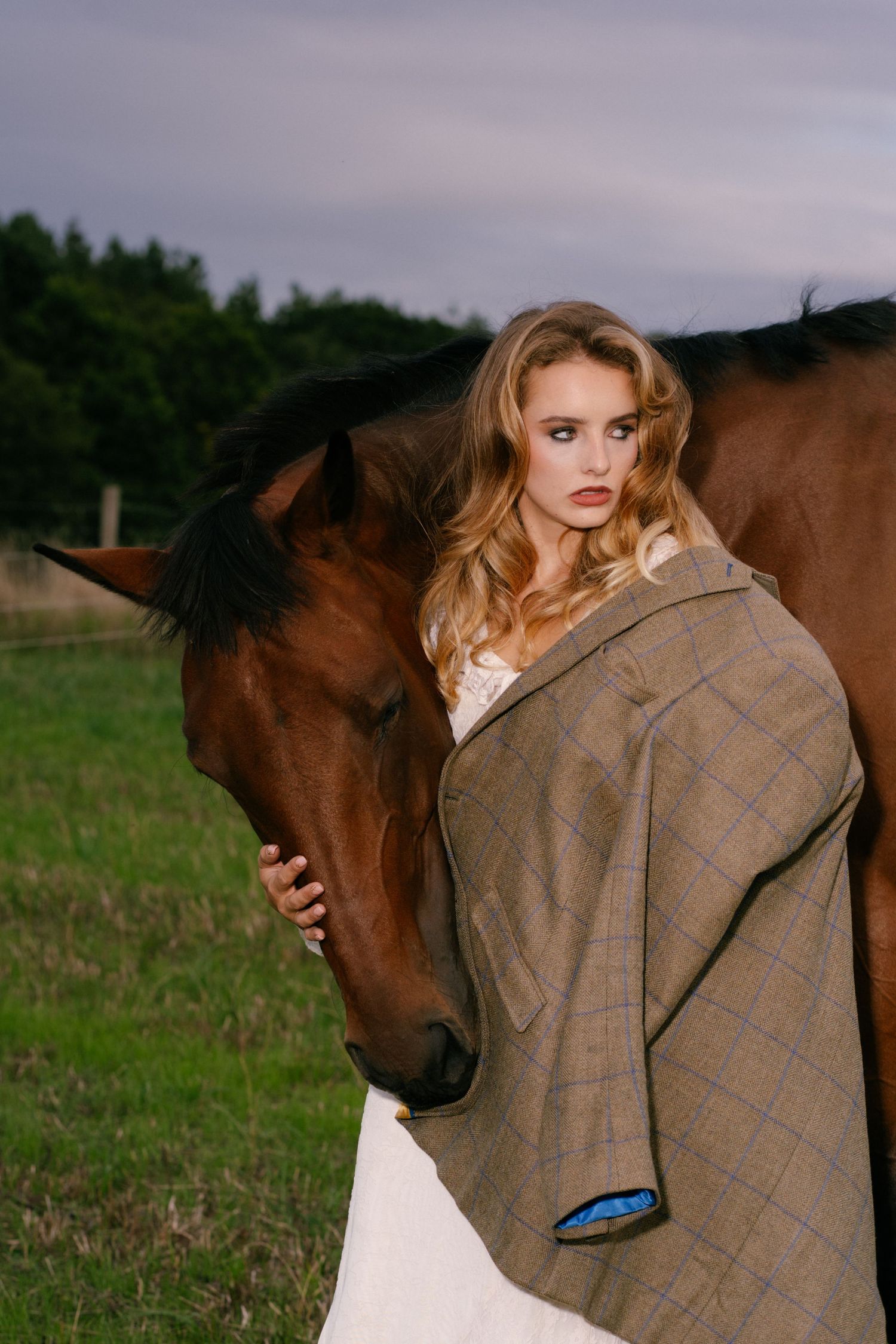 Blonde woman in plaid coat embracing a brown horse in a green field at dusk.