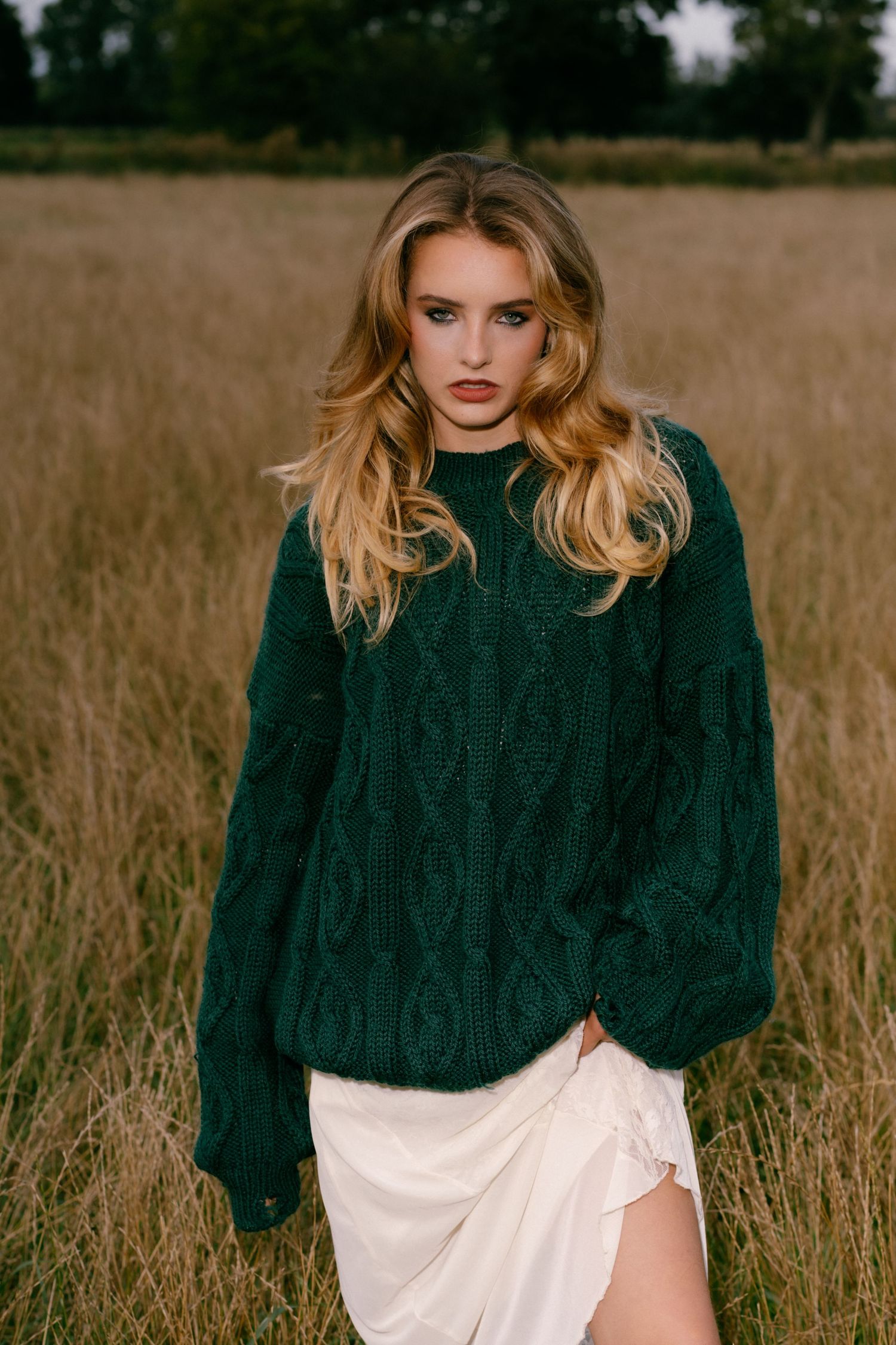 Young woman in green cable-knit sweater and white skirt standing in golden field with wavy blonde hair.