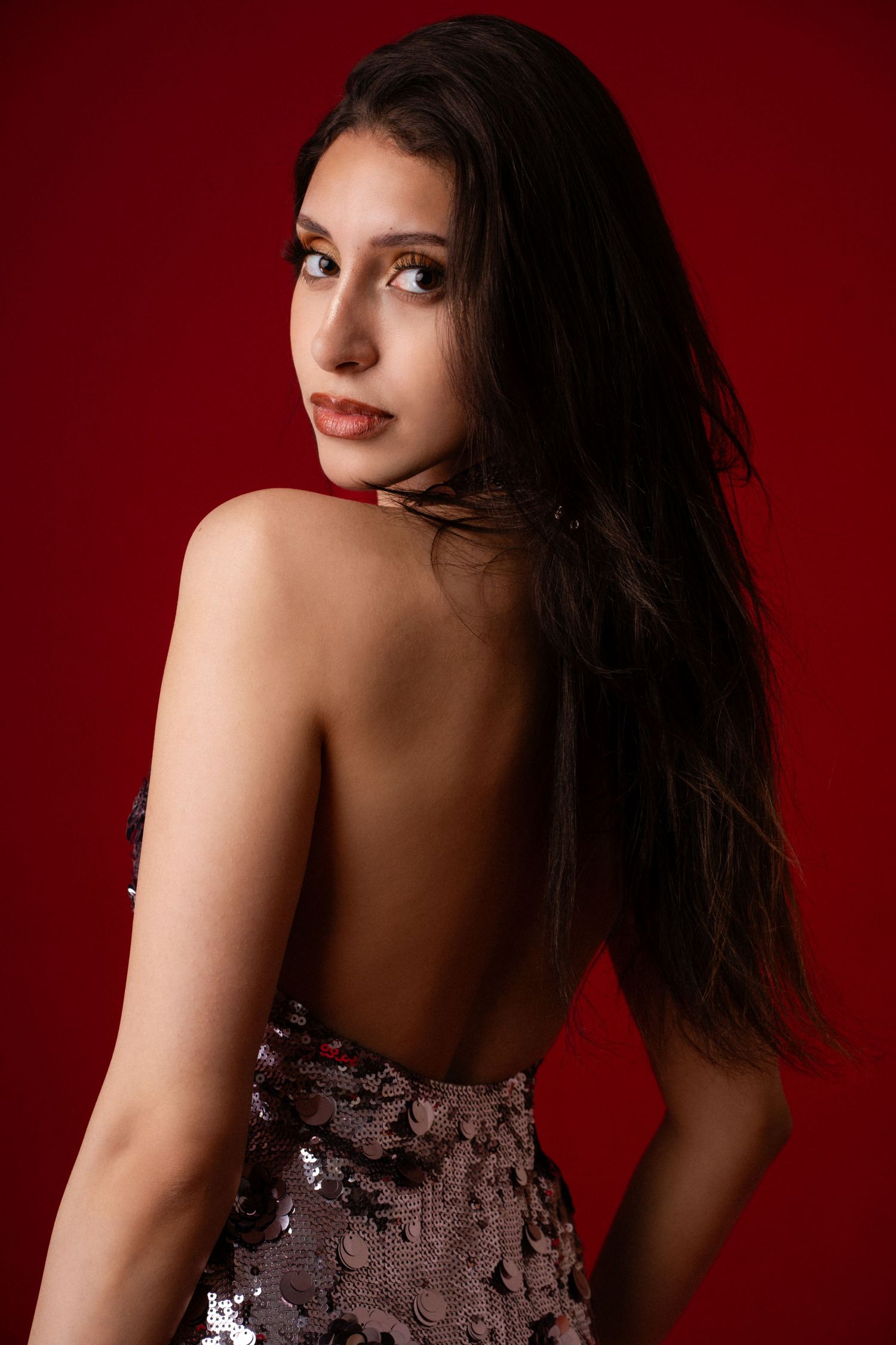 Young woman in backless sequin dress poses against red background, showing elegant bare back with long dark hair.