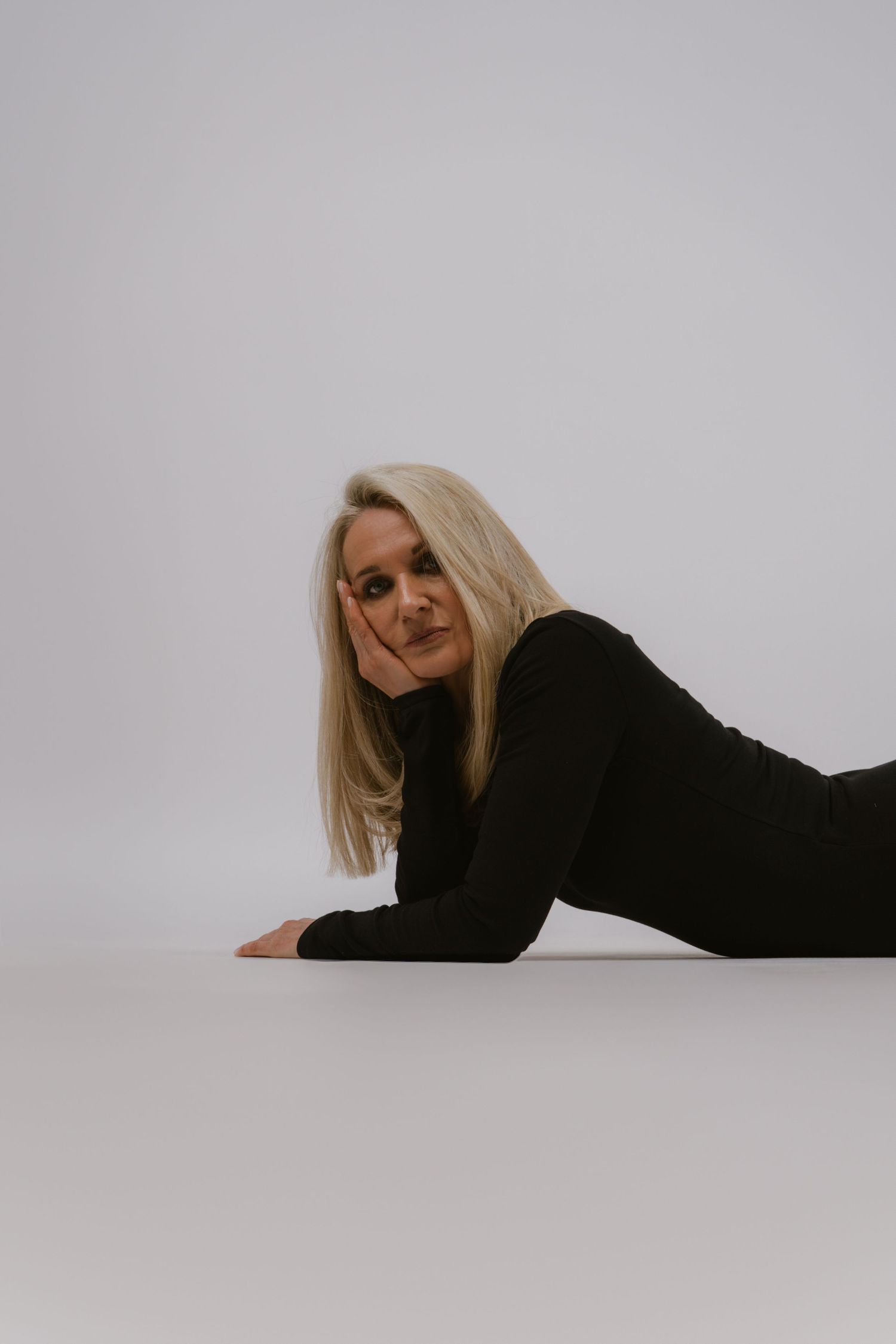 Blonde woman in black long-sleeve outfit lying on floor resting chin on hand in studio.