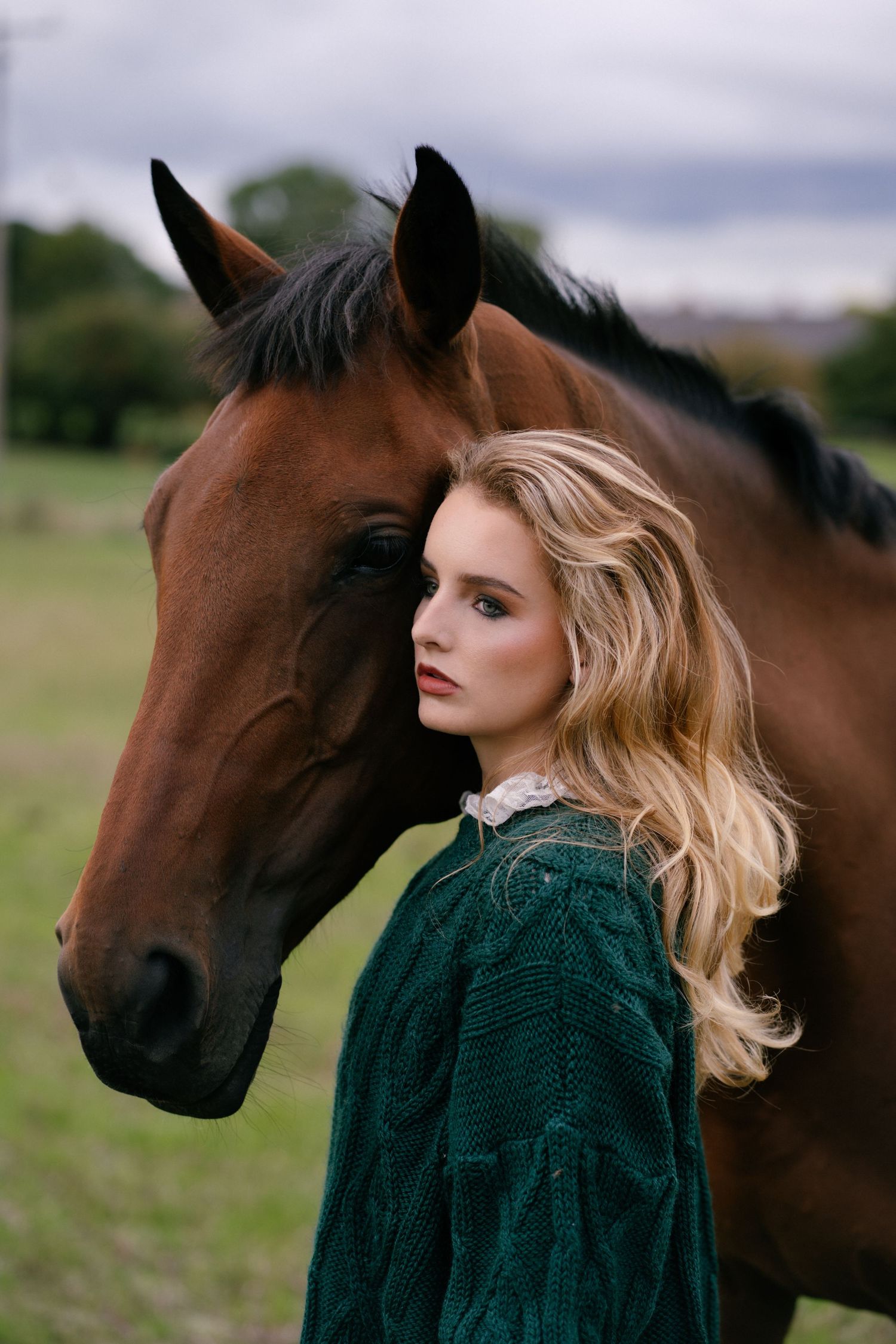 Young blonde woman in green dress poses with a bay horse in a field.