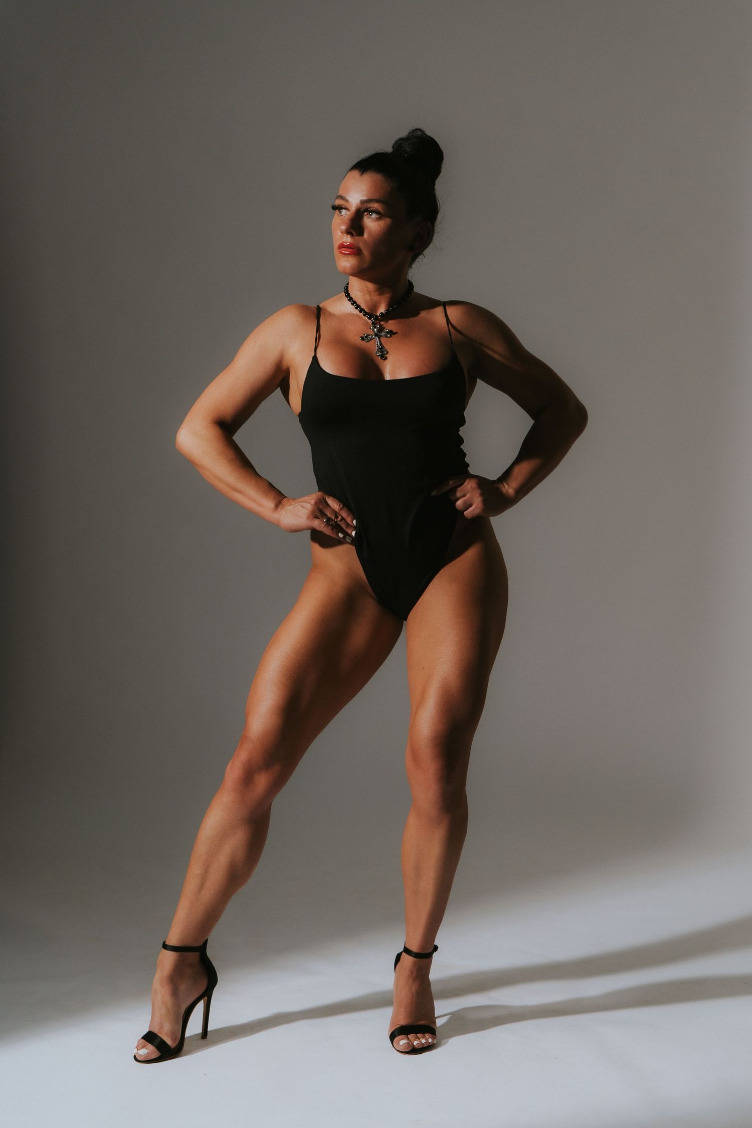 A toned woman in a black bodysuit with a statement necklace poses confidently with hands on hips in heels.