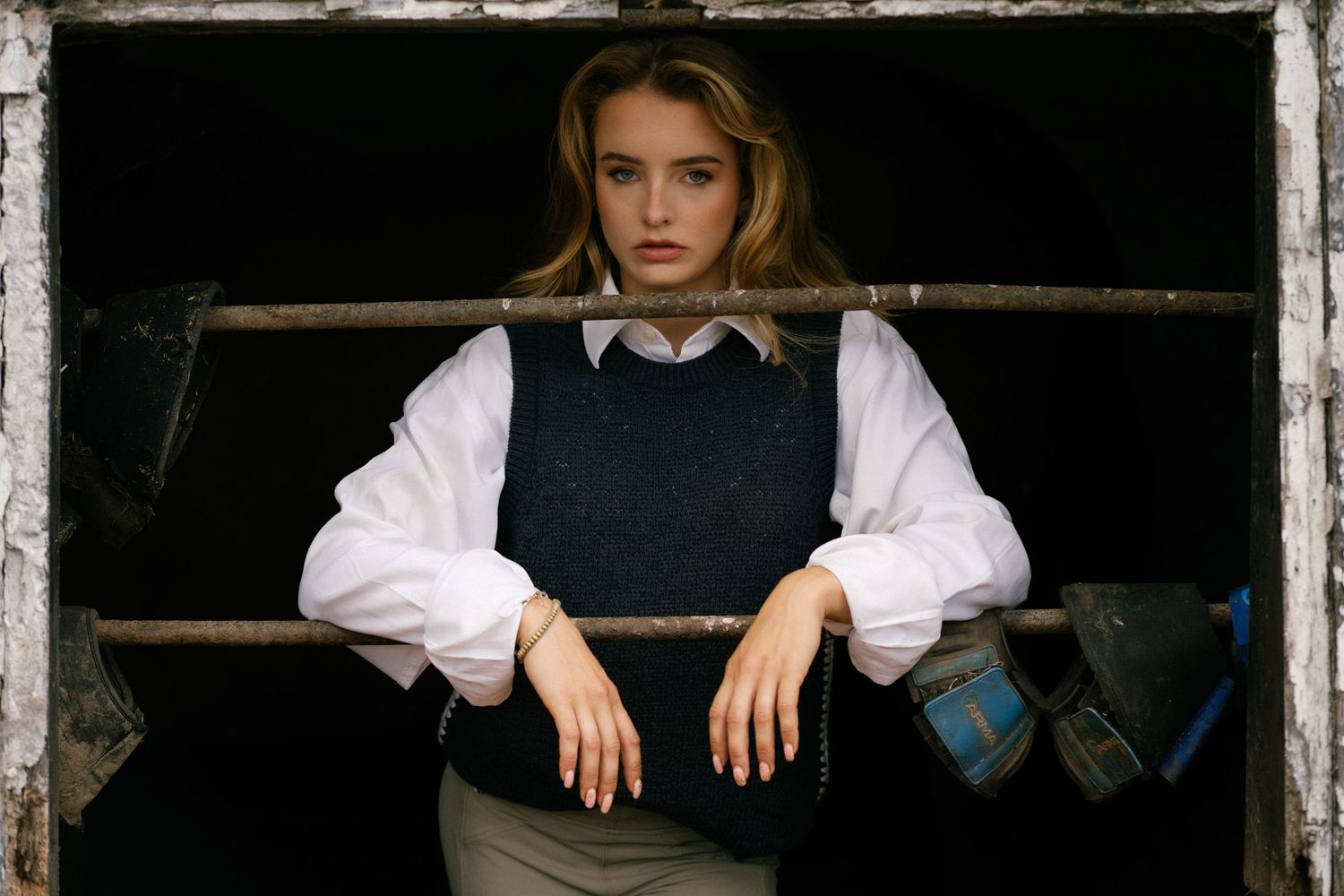 Young woman in white blouse and dark vest leans against rustic metal bars in a moody, dramatic portrait.
