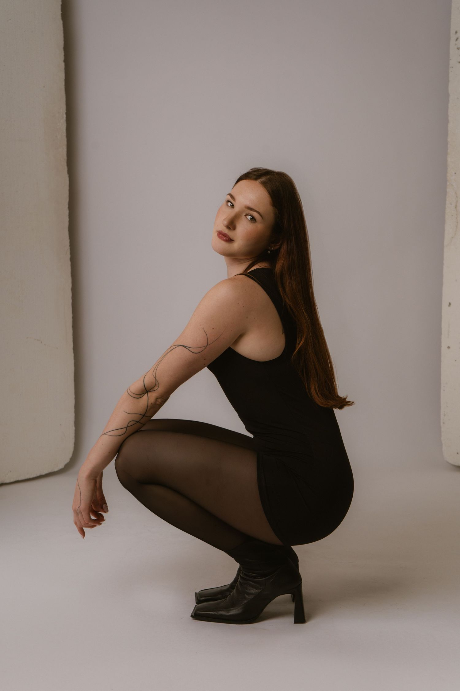 Woman in black bodysuit and tights crouching in studio, looking over shoulder, wearing black heeled boots.