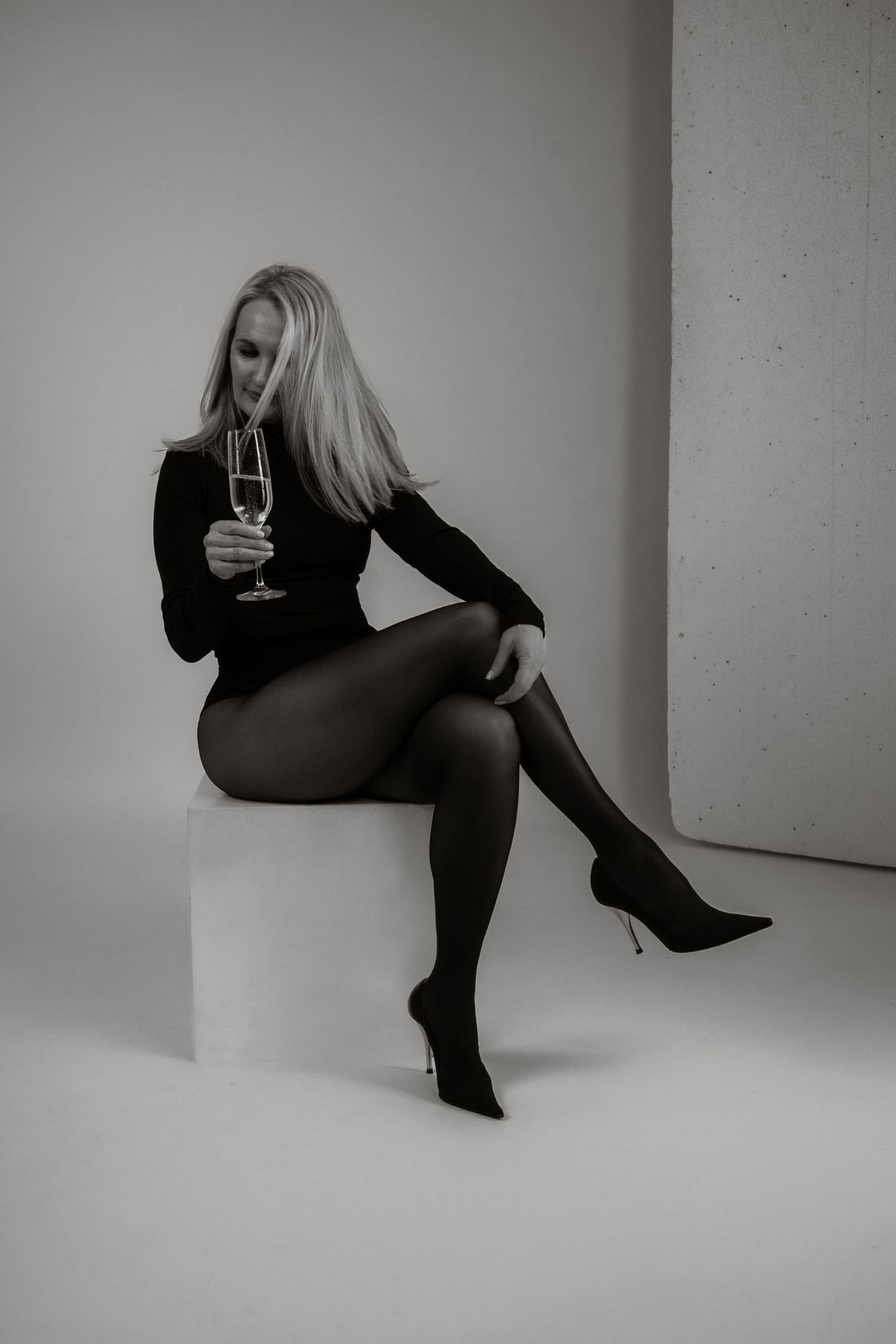 Black and white photo of blonde woman in black outfit holding champagne glass seated on cube.