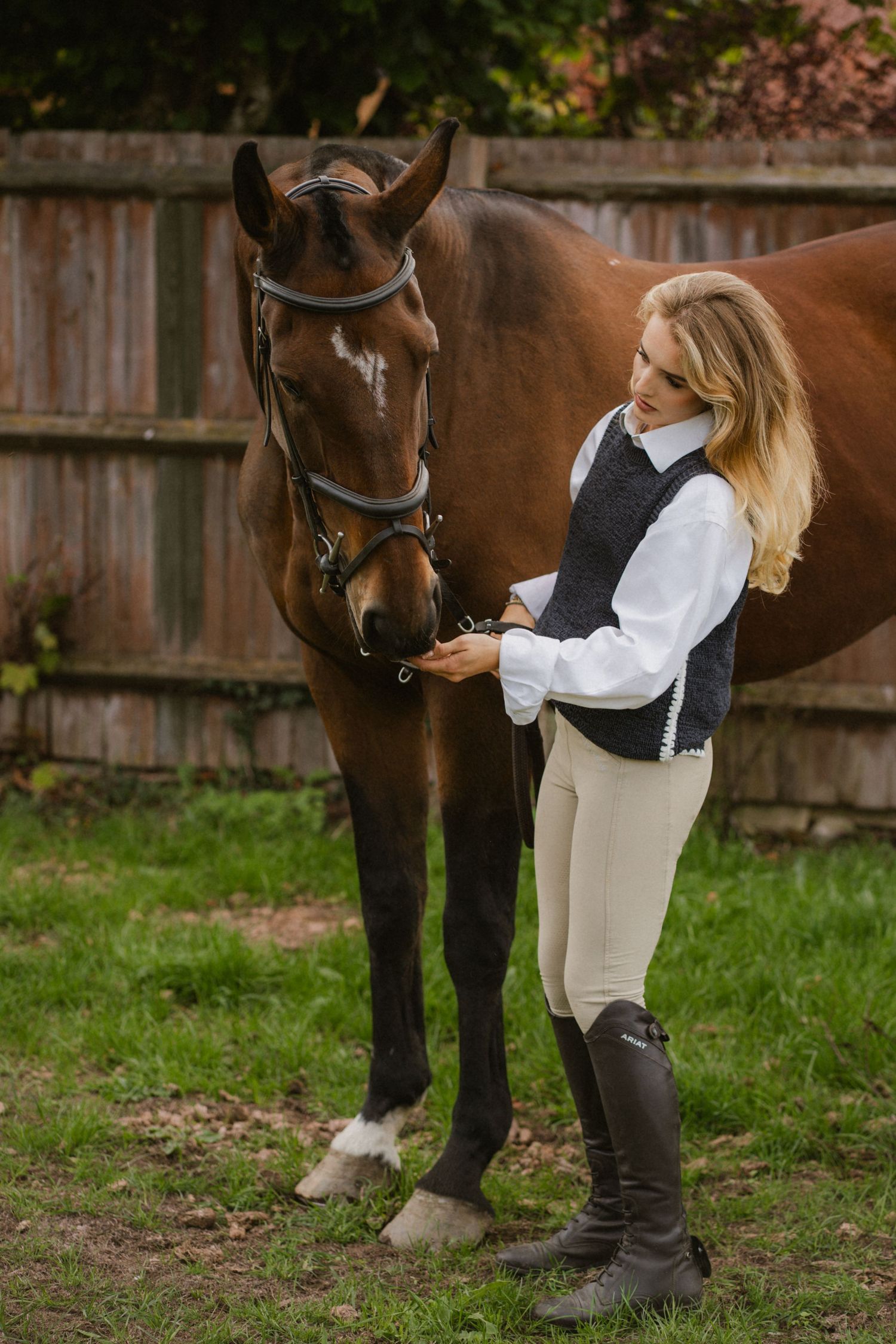 A blonde woman in equestrian attire stands beside a brown horse in a grassy outdoor setting.
