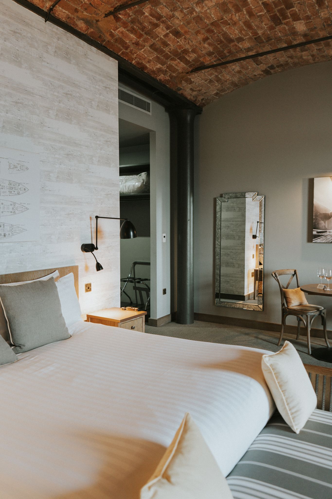 Modern hotel room with white bedding, exposed brick ceiling, full-length mirror, and wall sconce.