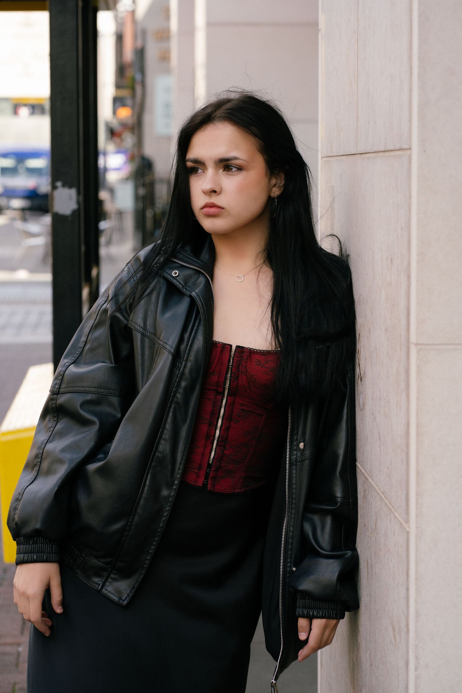 Young woman in black leather jacket and red corset top leaning against a wall on a city street.